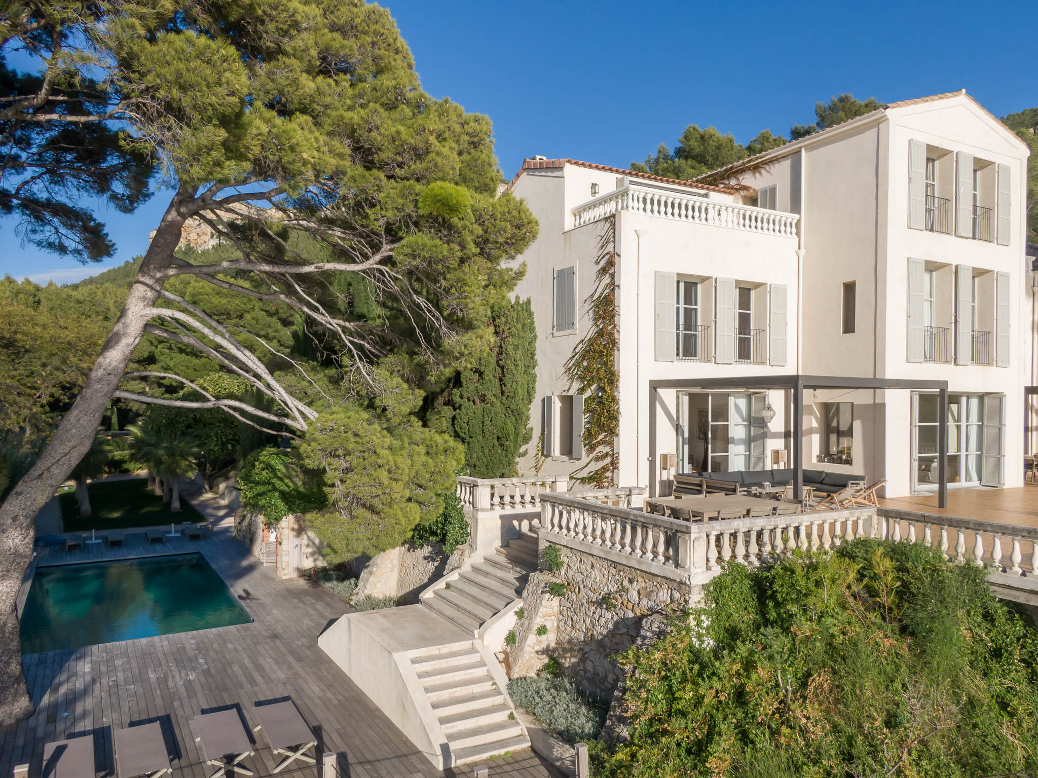 Aerial view of a white Provencal villa with grey shutters, terracotta roof, terrace seating, and swimming pool