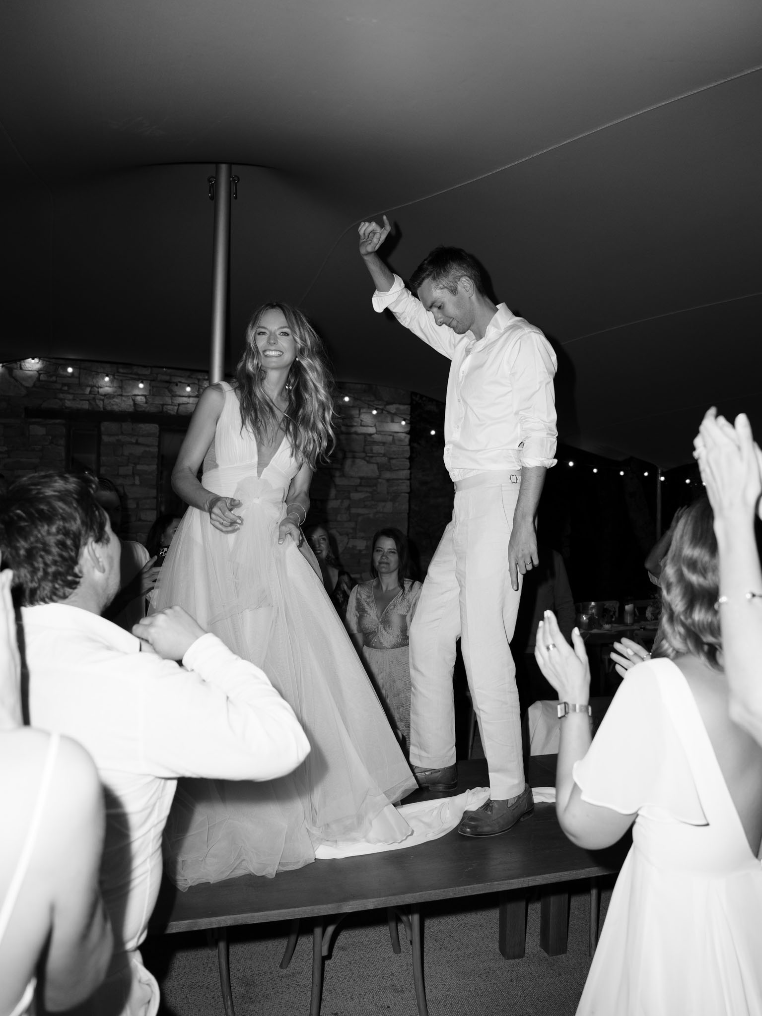 Black and white wide shot of couple dancing on table as guests cheer under tent with string lights