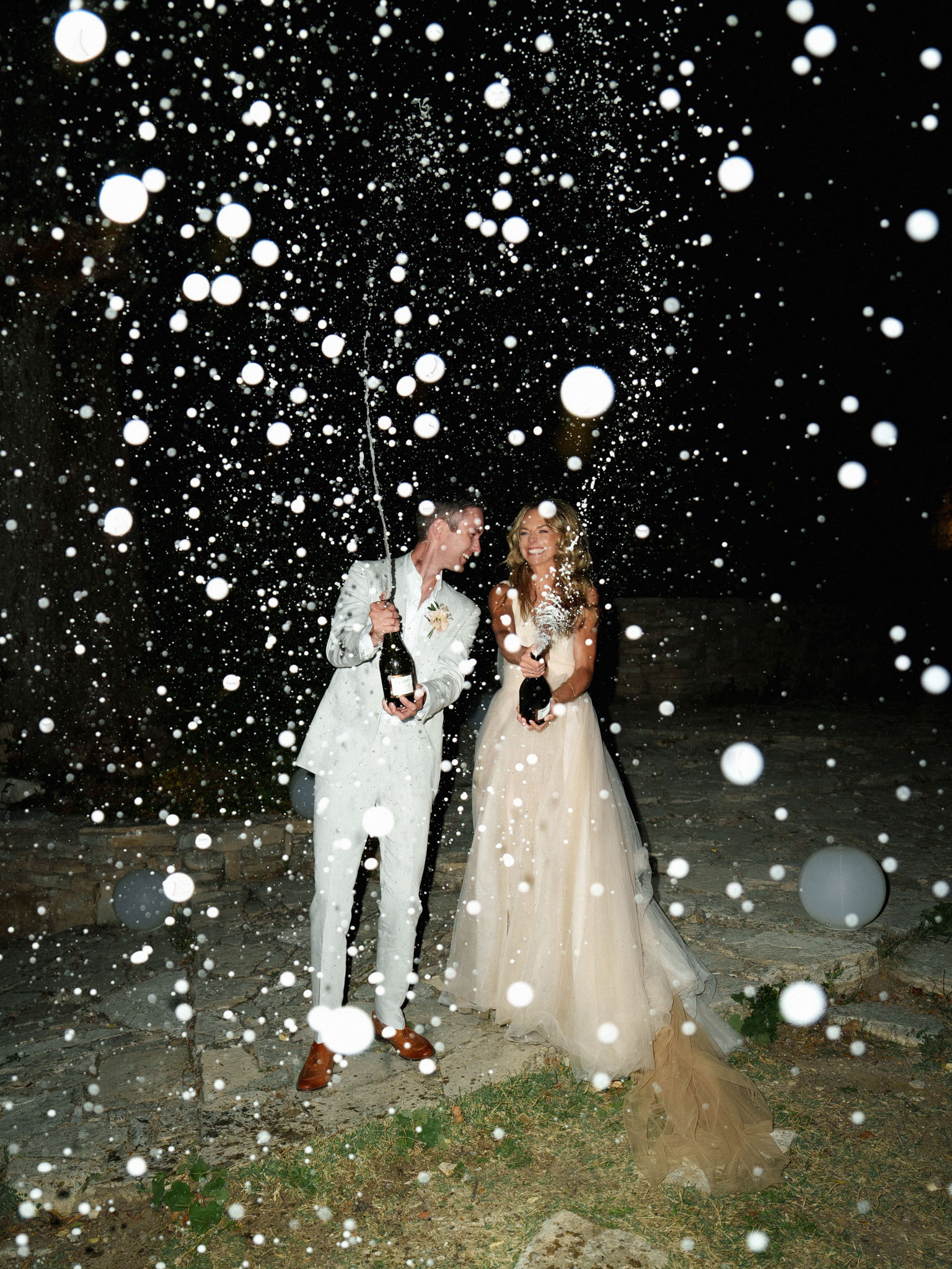 A bride and groom are photographed outdoors at night spraying champagne from two bottles, with droplets caught mid-air by the camera flash creating large circular bokeh orbs across the entire frame. The groom wears a light grey suit with brown leather shoes and a boutonniere, while the bride wears a champagne-toned layered tulle ballgown and holds a small white bouquet. Both are laughing and facing each other on a stone-paved outdoor area, with grey balloons visible on the ground nearby. The image is a full-length portrait shot with dramatic nighttime contrast, the flash illuminating the couple and champagne spray against a dark background.