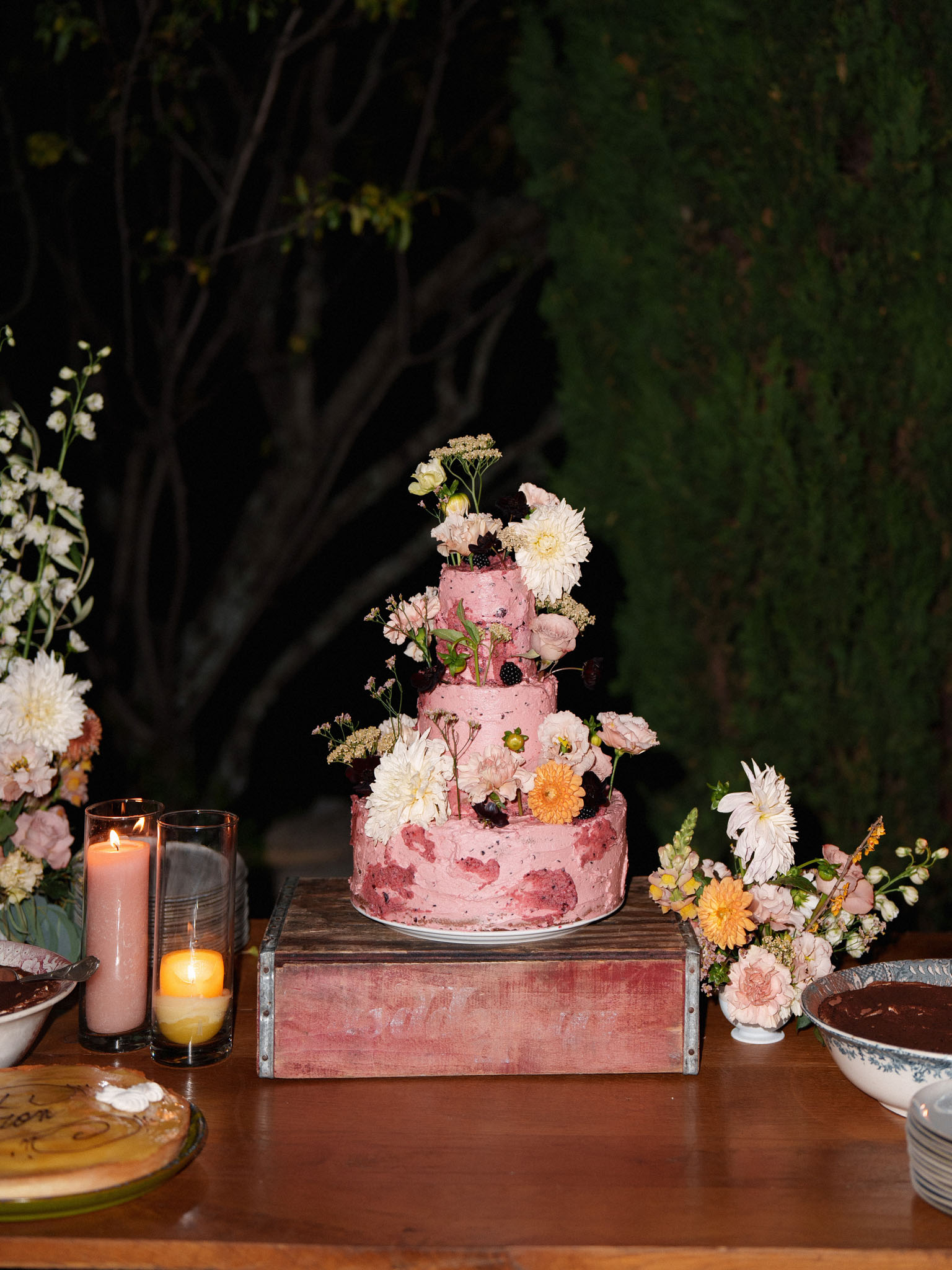Three-tier dusty rose cake with cascading dahlias, blackberries, and orange gerberas on wooden crate