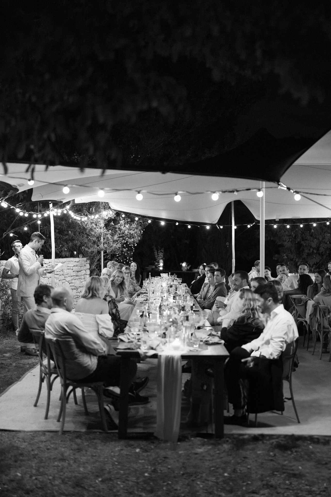 This black-and-white image shows an outdoor evening wedding reception dinner with approximately 20–25 guests seated along a single long banquet-style table under a stretched canopy tent. String globe lights are strung beneath the tent canopy, providing warm, glowing illumination against the dark night sky. The table is set with glassware, candles, and a fabric table runner running its full length, with place settings and small floral or decorative centerpieces visible in mid-tones. Guests are dressed in smart-casual to semi-formal attire, engaged in conversation. Two guests stand at the far left near a low stone wall, holding drinks. The overall styling suggests a relaxed yet dressed outdoor reception, likely at a French countryside property with rustic stone boundary walls visible in the background. The shot is a medium-wide perspective taken from one end of the table, capturing the full length of the dining setup and the tent structure above.