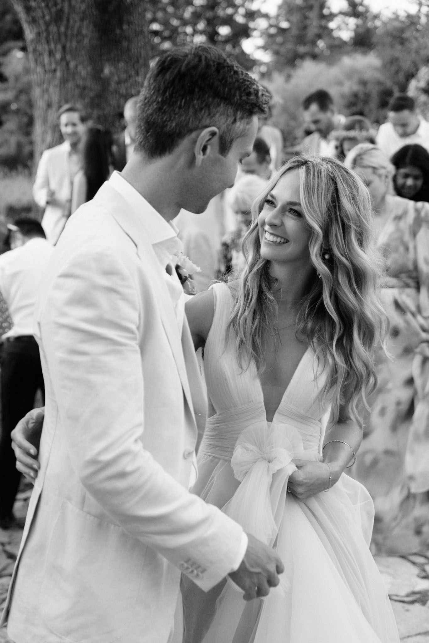 Black and white portrait of laughing bride and groom holding hands outdoors with guests blurred in background