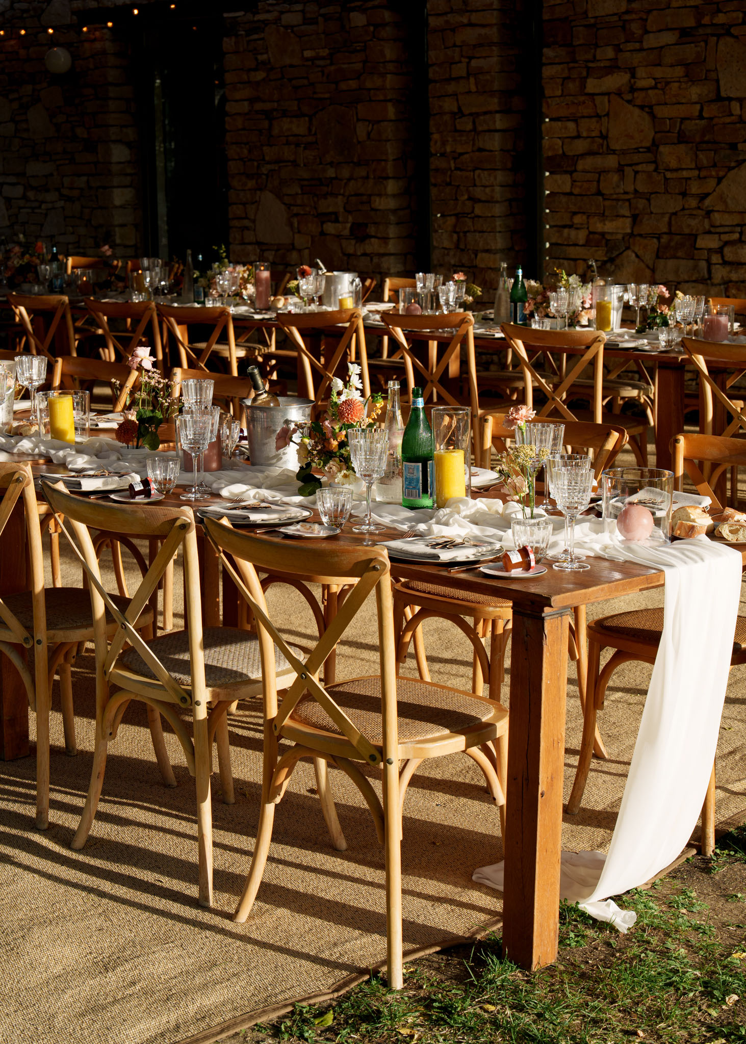 Outdoor reception tables under stone-walled structure with coral dahlia centerpieces, pillar candles, and cross-back chairs