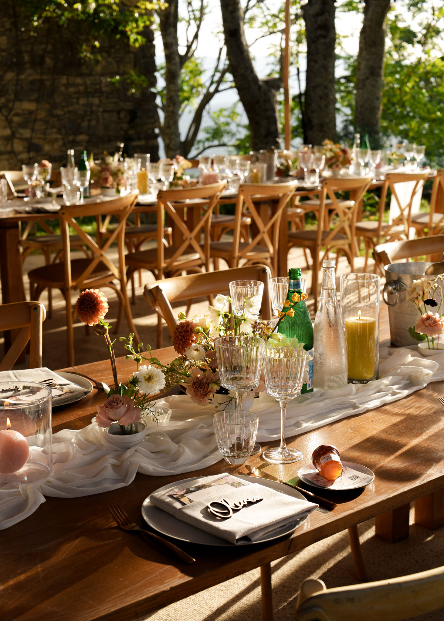 Farm table with terracotta dahlias, yellow candles, and gold cutlery on gauze runner under trees