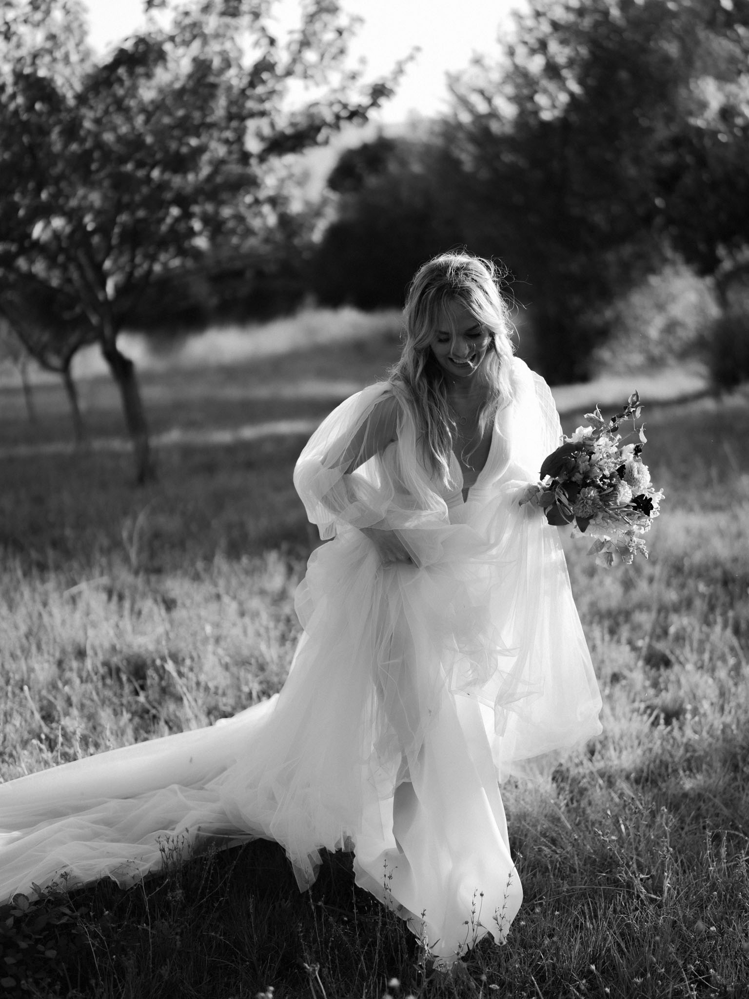 Black-and-white portrait of bride laughing in puffed-sleeve tulle ball gown holding mixed bouquet in orchard setting