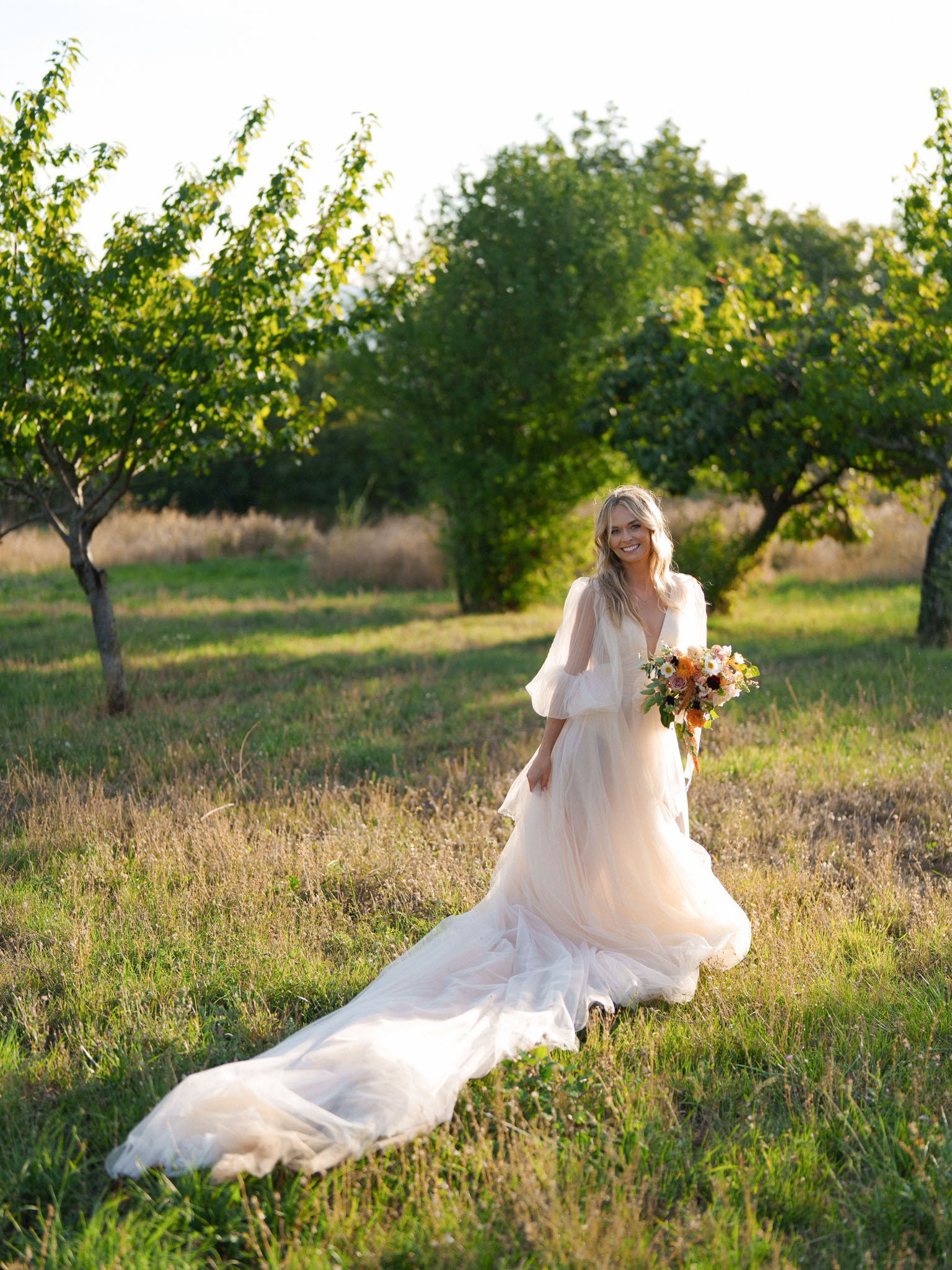 A bridal portrait taken outdoors in an orchard or meadow setting during golden hour, with warm low-angle sunlight illuminating the scene. The bride, a blonde woman with loose wavy hair, walks toward the camera smiling, wearing a blush-toned tulle gown with a deep V-neckline, billowy three-quarter sleeves, and an extended cathedral-length train spread across the grass. She holds a loose, garden-style bouquet in warm tones of burnt orange, coral, peach, and ivory, featuring roses, dahlias, and small daisy-like blooms with trailing ribbons. The overall styling is boho-romantic, and the shot is a full-length portrait with the train dramatically extending into the foreground.