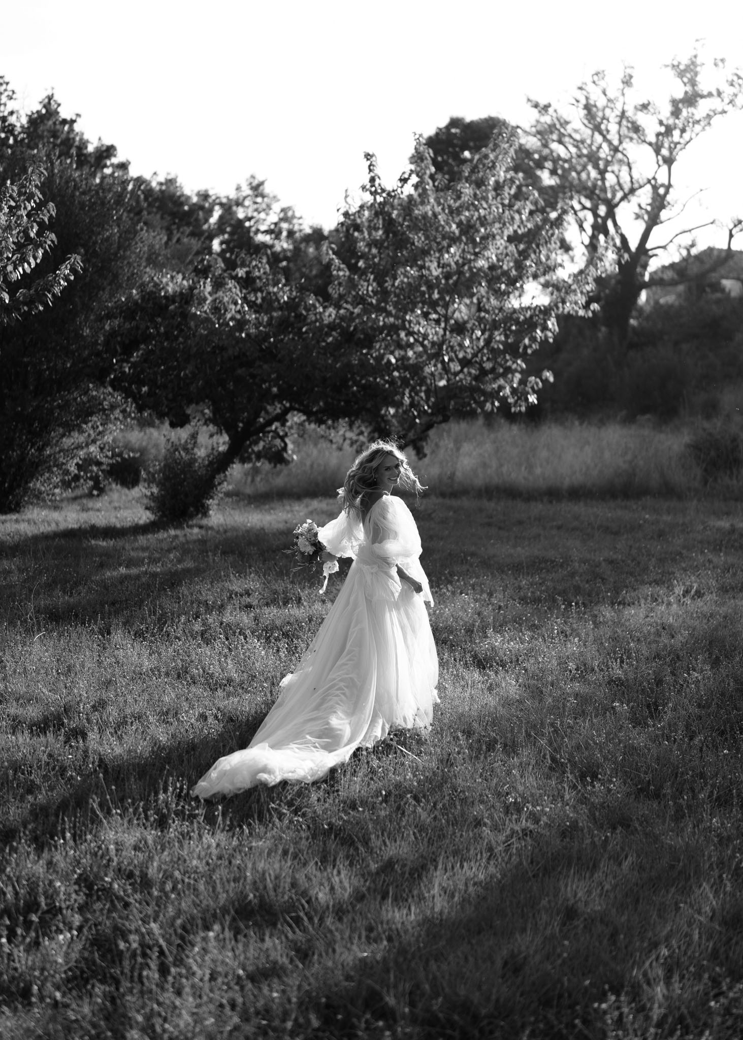 Black-and-white portrait of bride walking through a field with flowing gown and long train