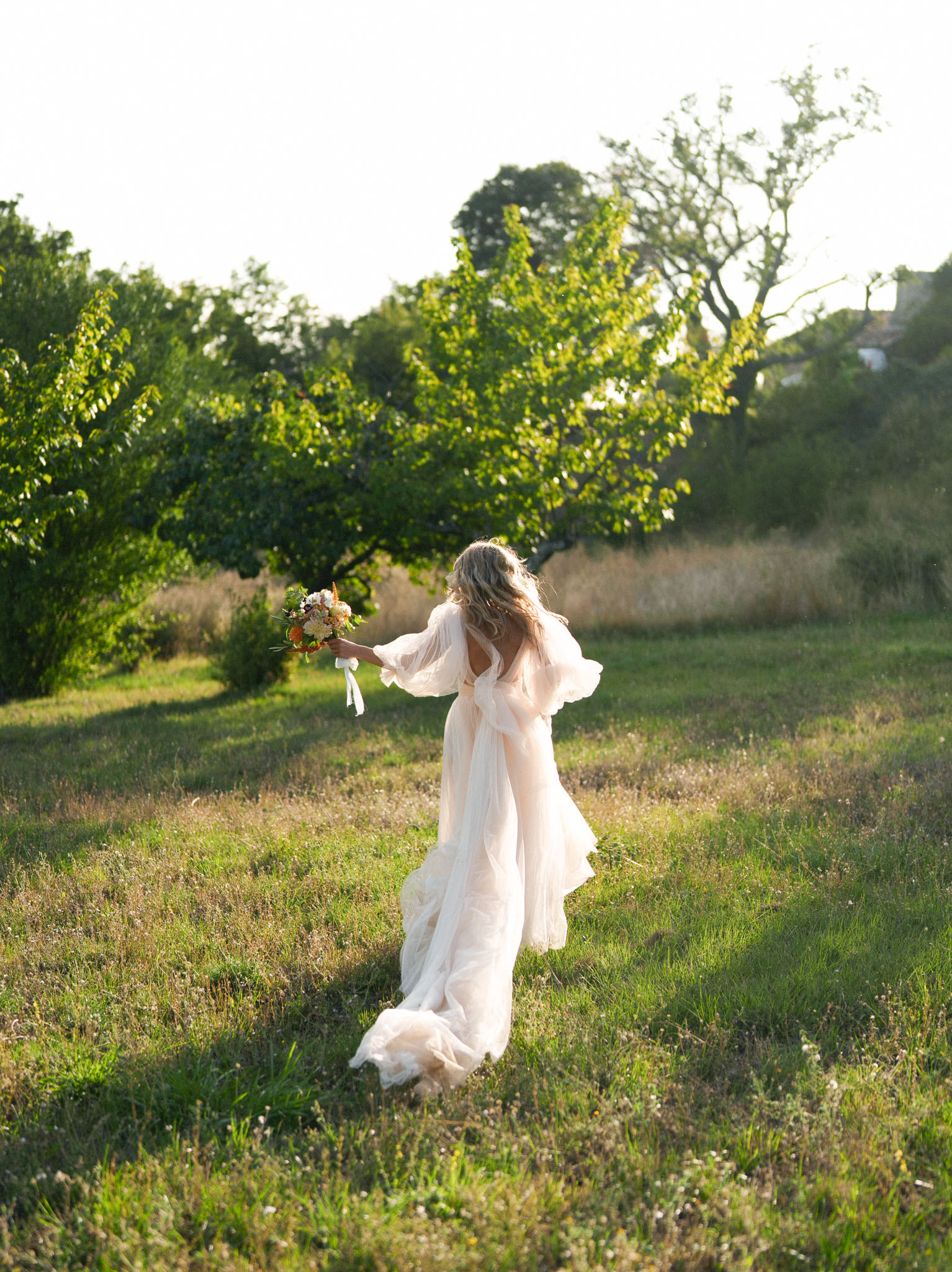 Bride walking through orchard at golden hour in flowing chiffon gown holding garden-style bouquet