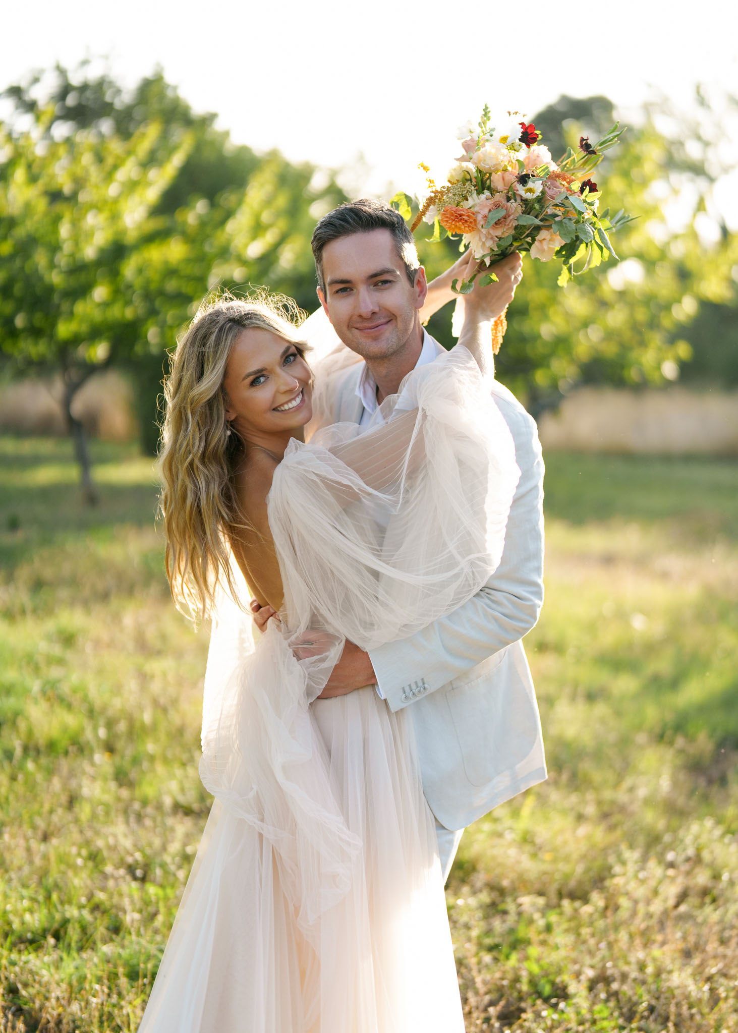 Couple embracing in orchard at golden hour, groom holding blush and burgundy bouquet