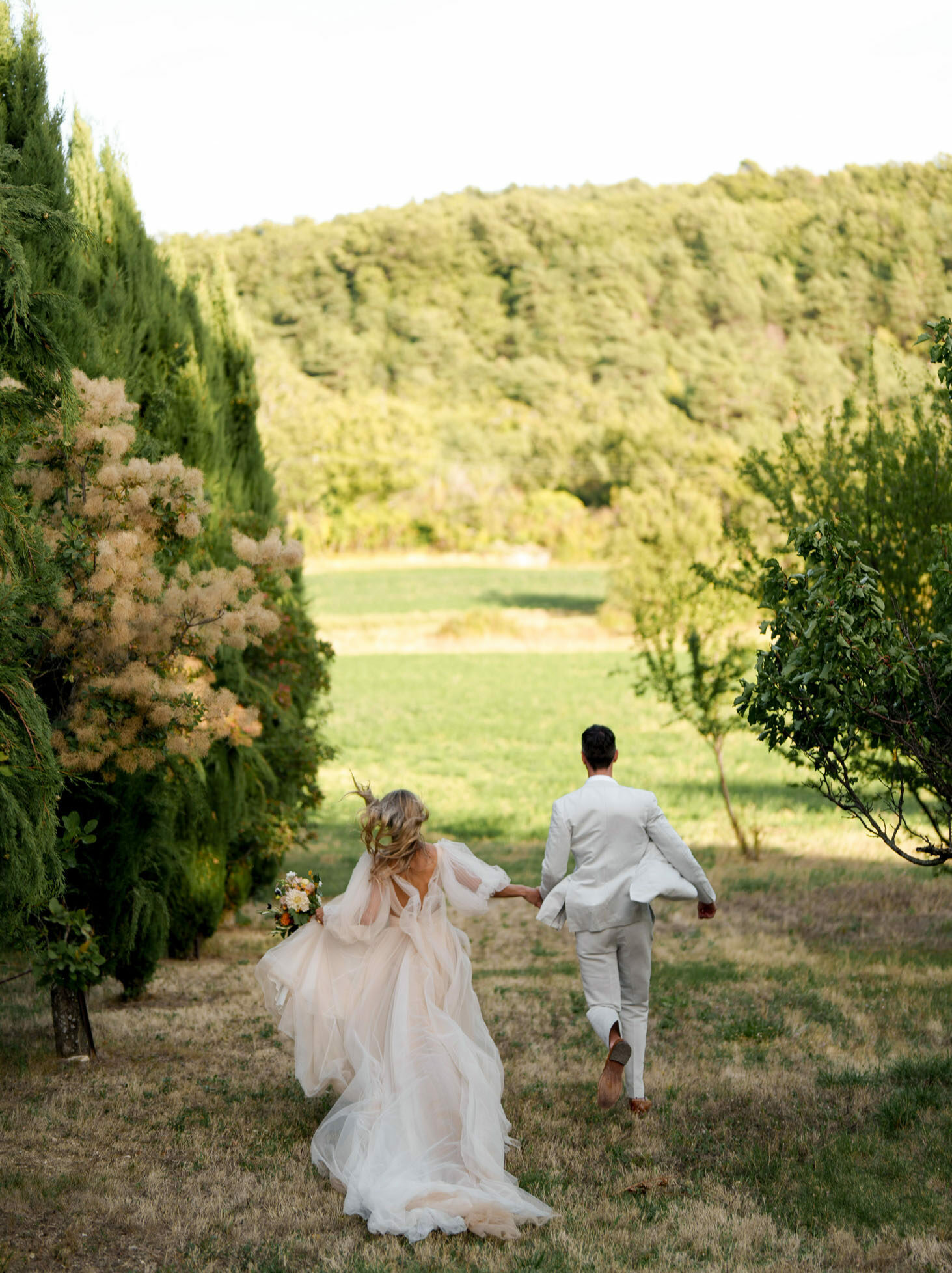 Couple running hand in hand through tree-lined path in blush tulle gown and grey suit