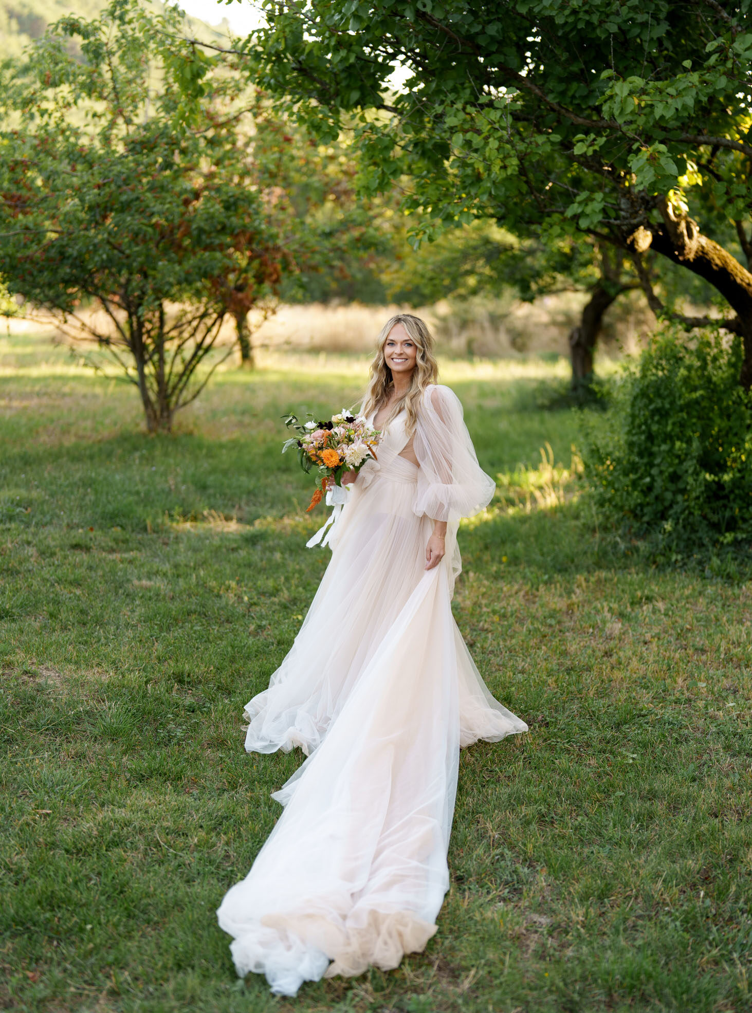 A bridal portrait taken outdoors in what appears to be an orchard or garden setting during golden hour. The bride stands alone, smiling at the camera, wearing a blush-toned tulle A-line gown with voluminous puff sleeves, a V-neckline, and a long flowing train. She holds a loose, organic-style bouquet featuring orange dahlias, blush blooms, dark burgundy anemones, and greenery with trailing white ribbon streamers. Her wavy blonde hair is worn loose. The overall styling is romantic and boho in feel. Full-length portrait shot with a shallow depth of field.