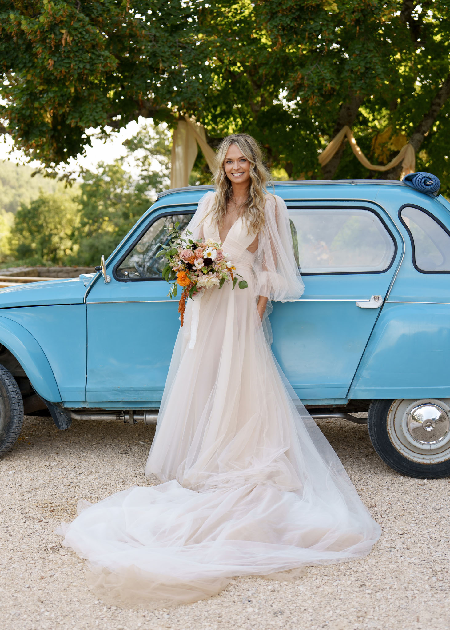 A bride stands outdoors on a gravel surface in front of a vintage light blue Citroën 2CV, posing for a full-length portrait. She wears an ivory tulle A-line gown with a deep V-neckline, voluminous sheer puff sleeves, and an extended cathedral-length train spread across the ground. Her loose, wavy blonde hair is worn down, and she holds a lush bouquet featuring blush and mauve roses, white daisy-like blooms, deep burgundy flowers, orange marigolds or amaranth, and trailing greenery. The overall styling has a relaxed boho sensibility, with the vintage car serving as a deliberate decorative prop. The image is a mid-length to full portrait shot taken in natural daylight.