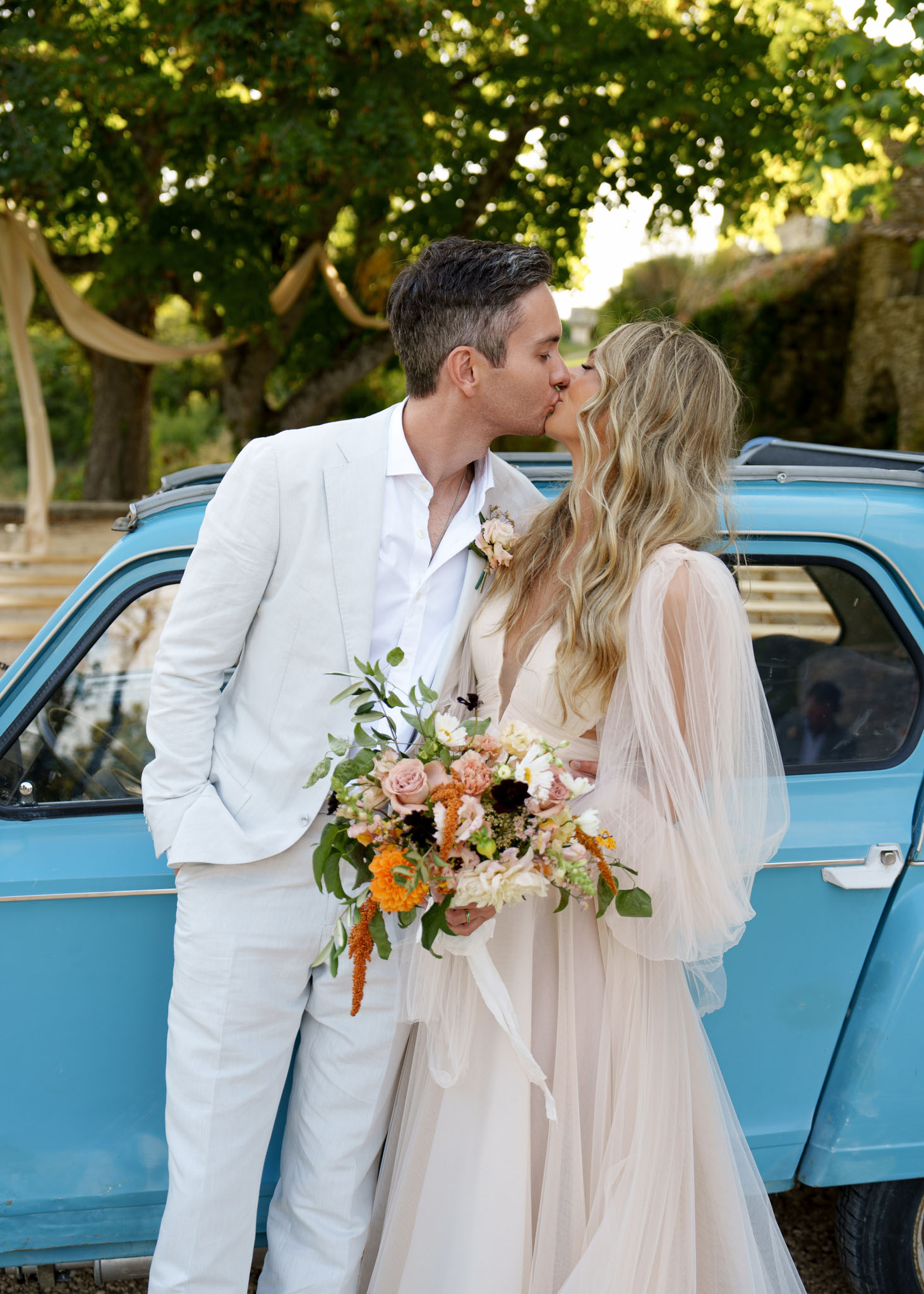 A couple portrait taken outdoors, with the bride and groom kissing in front of a vintage powder-blue Citroën 2CV or similar retro French car. The groom wears a light cream linen suit with an open-collar white shirt and a small blush floral boutonniere. The bride wears a blush-toned gown with a deep V-neckline and flowing sheer tulle sleeves, with long wavy blonde hair worn down. She holds a large loose bouquet featuring blush and mauve roses, deep burgundy anemones, orange marigolds, trailing amaranthus in rust tones, white blooms, and abundant greenery with long white ribbon streamers. The overall styling is boho with a warm autumnal color palette. The shot is a mid-length portrait framed tightly on the couple with the car providing a colorful backdrop.