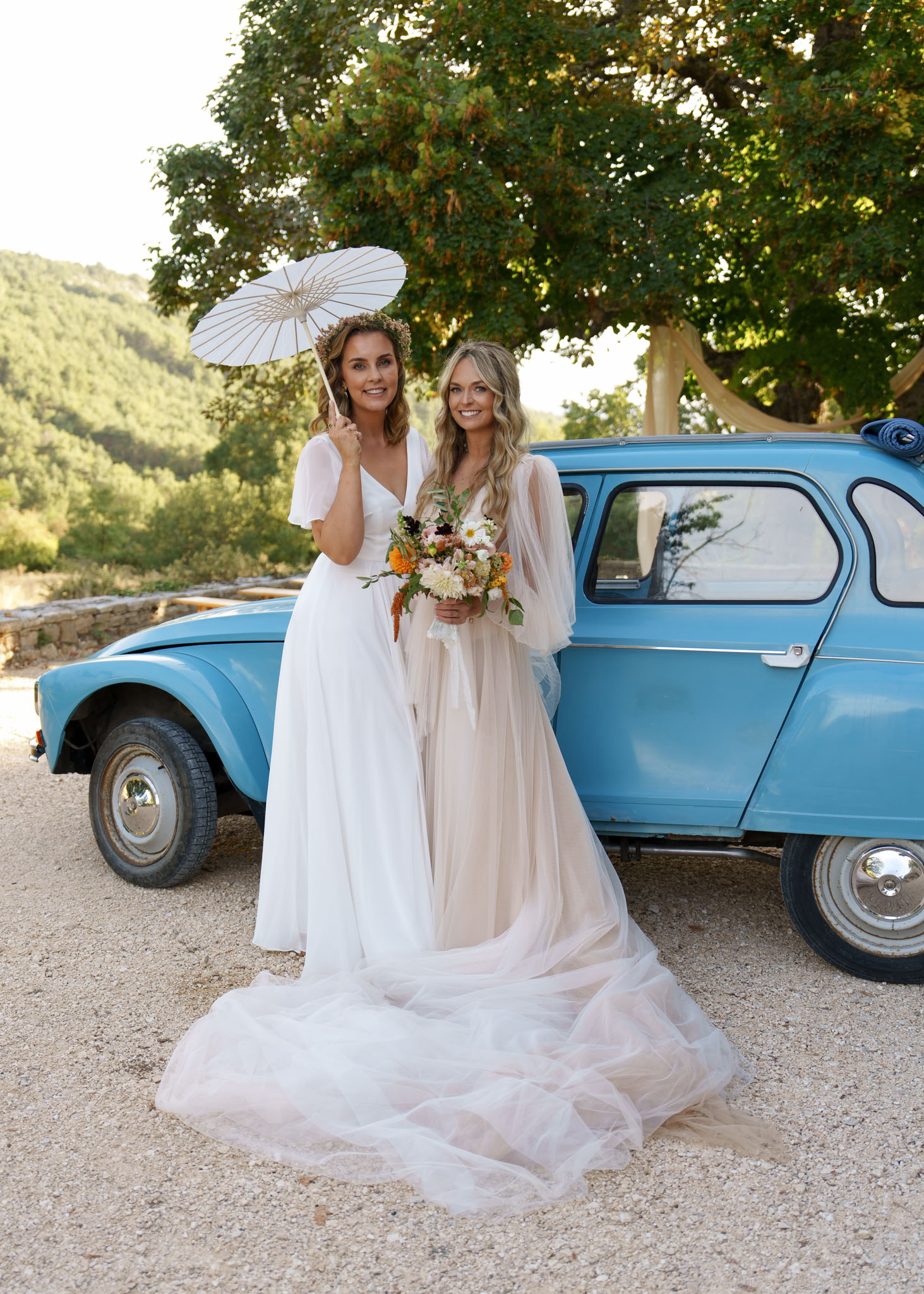 Two women in boho gowns with floral crown and dahlia bouquet pose beside vintage baby-blue Renault 4