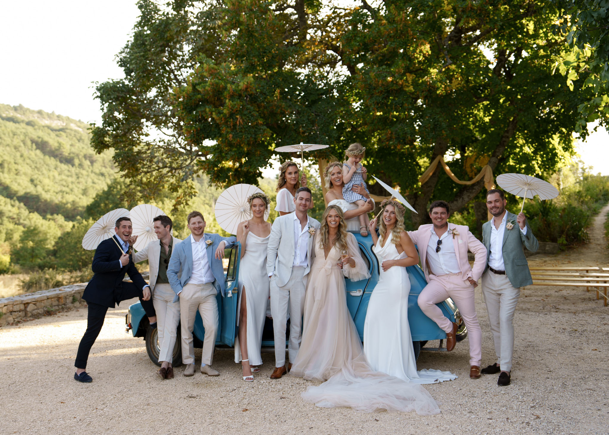 Bridal party group portrait around vintage blue Citroen 2CV on gravel driveway with paper parasols