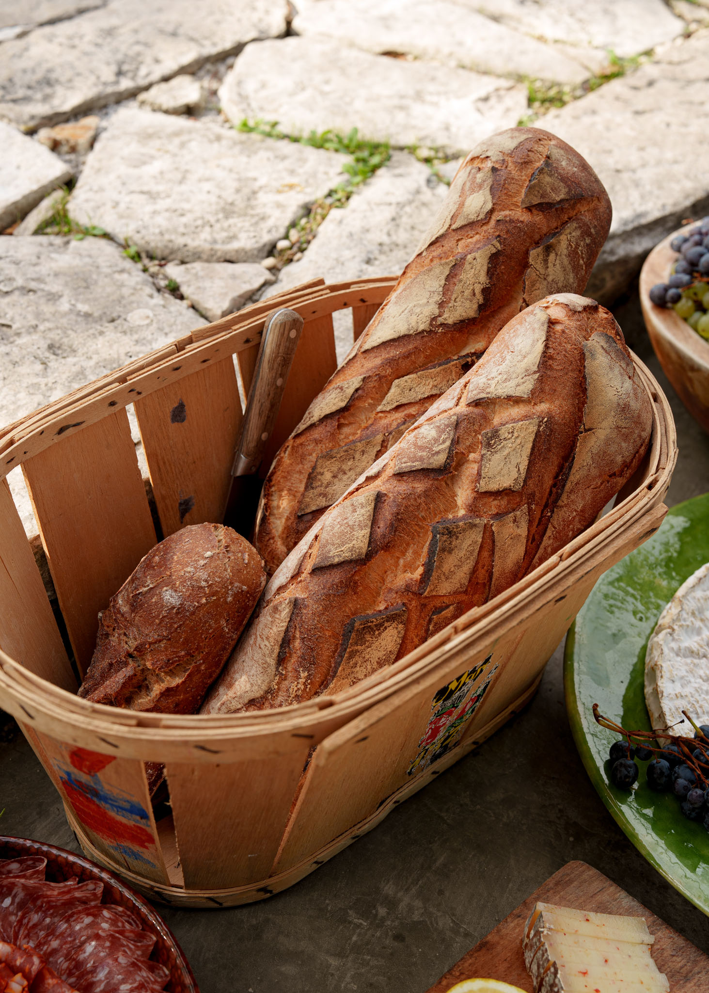 Rustic bread basket with baguettes surrounded by brie, grapes, and cured meats on stone surface