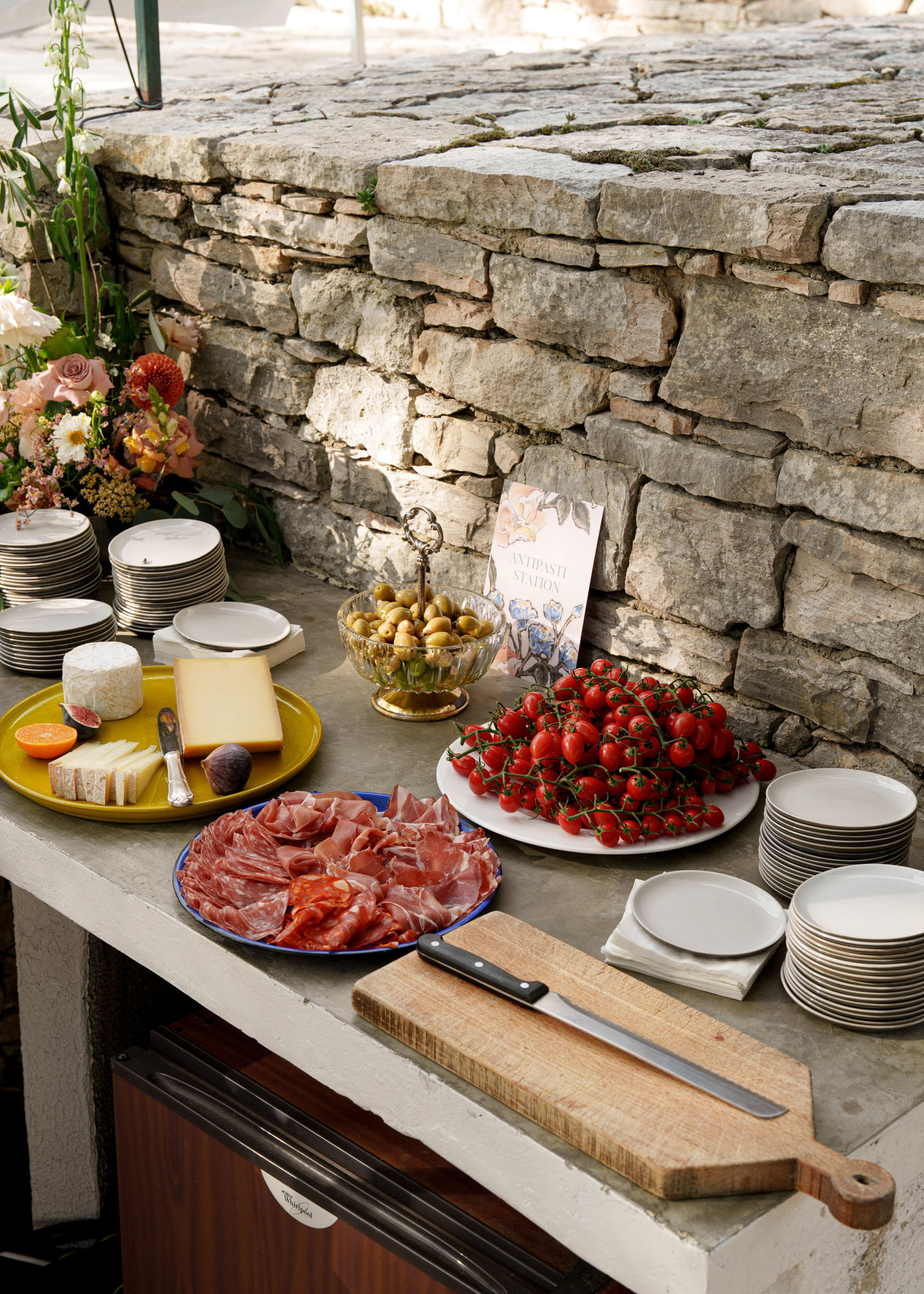 Antipasti station with cheeses, charcuterie, tomatoes, and olives against a stone wall at a countryside venue