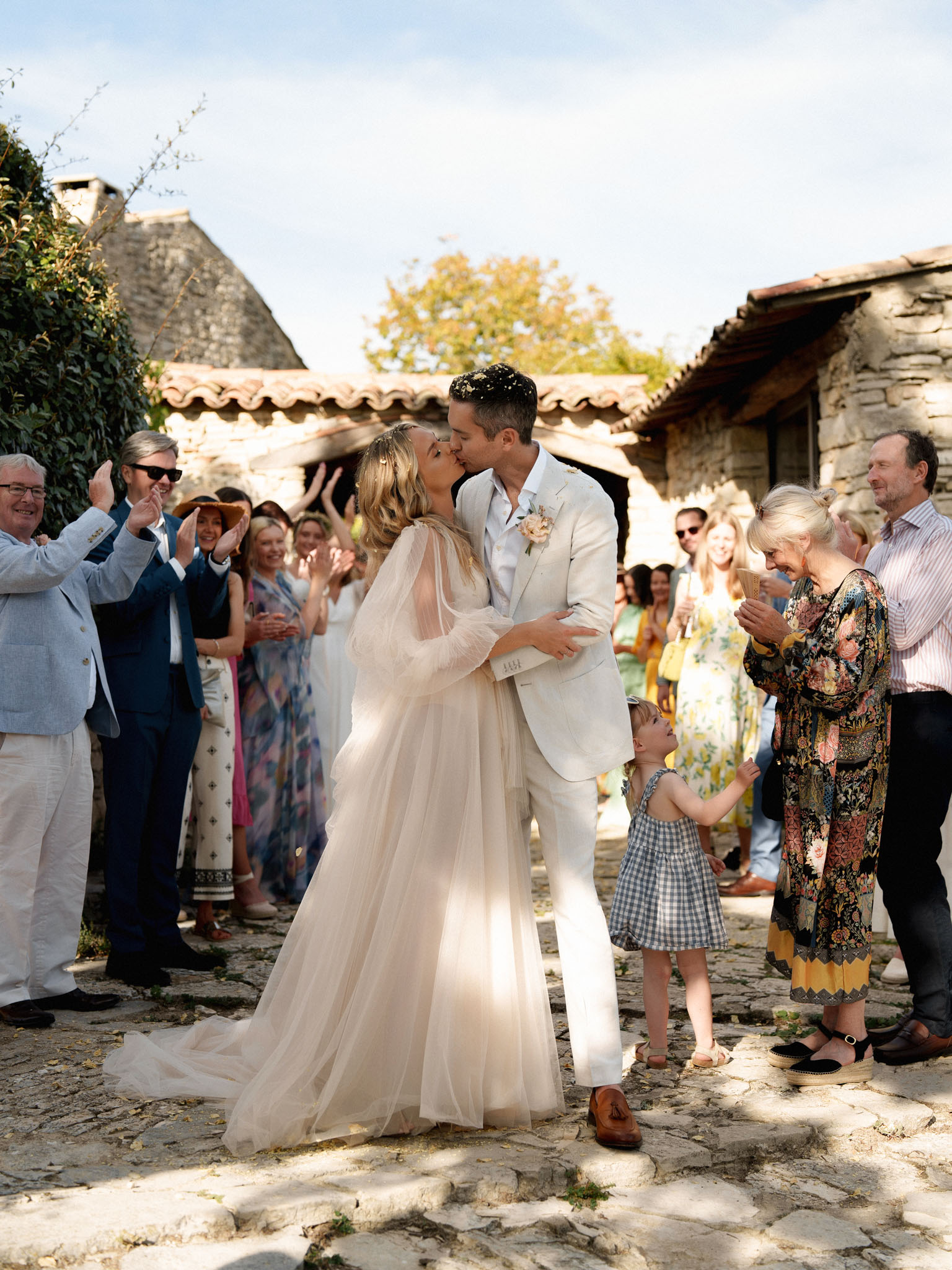 The couple shares a first kiss on a cobblestone courtyard surrounded by approximately 20 applauding guests lining both sides to form a celebratory aisle. The bride wears a soft blush-toned tulle gown with long sheer sleeves and a flowing skirt, paired with a sheer tulle veil and a floral hair accessory; small flower petals are visible in the air around the couple. The groom wears an off-white linen suit with a pale pink boutonnière. The setting is an outdoor rustic French stone farmhouse courtyard with terracotta-tiled rooflines and cobblestone paving, consistent with a Provence or Ardèche-style property. Guests are dressed in colorful summer attire, and a young child in a blue gingham dress stands nearby watching; the overall styling aesthetic is relaxed and romantic with a natural, sun-lit daytime atmosphere. Wide portrait shot with the couple centered in the foreground.