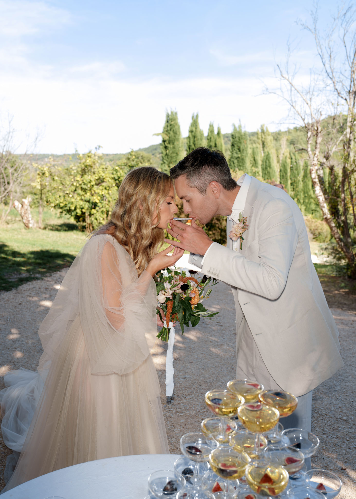 Bride and groom toasting with champagne coupes during outdoor cocktail hour with champagne tower on table