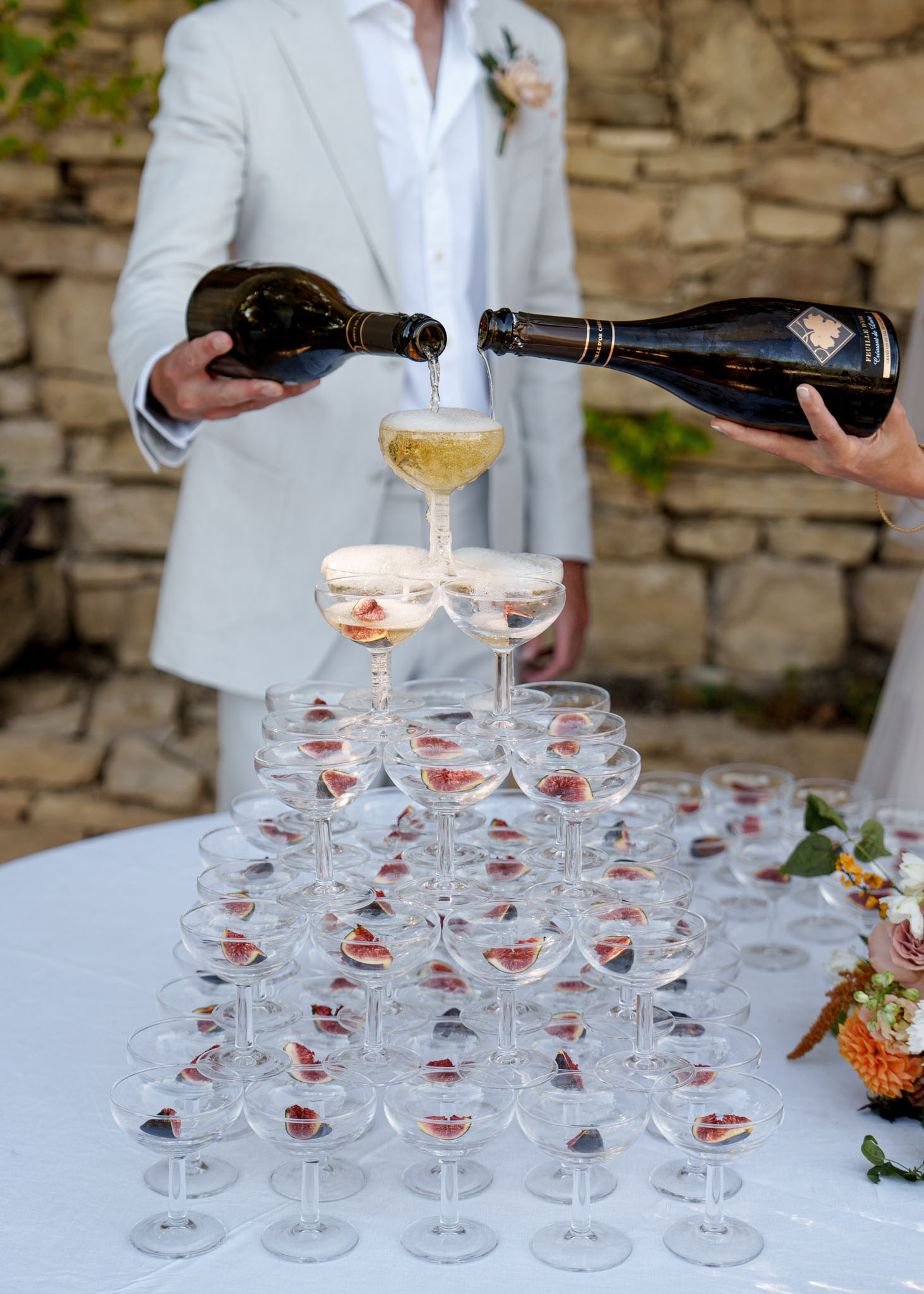 Champagne tower pouring with fresh fig halves in each coupe glass beside orange dahlia arrangement