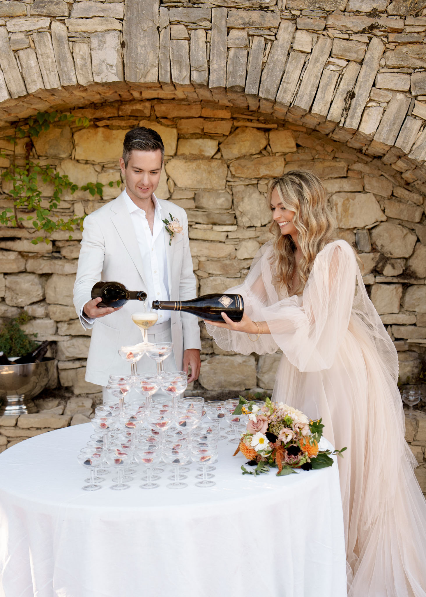 Bride in blush tulle gown and groom in ivory suit pouring champagne into coupe glass tower outdoors