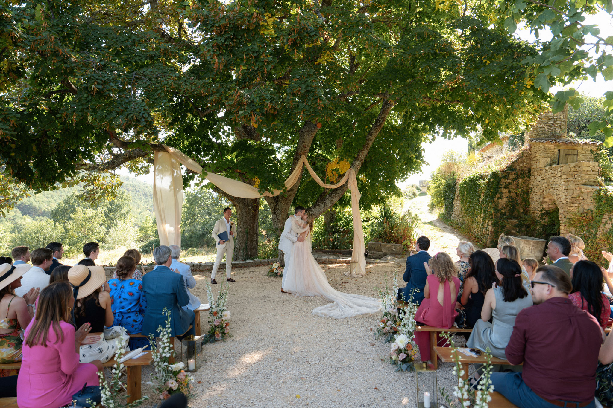First kiss under tree altar with draped cream fabric and blush floral aisle on rustic terrace