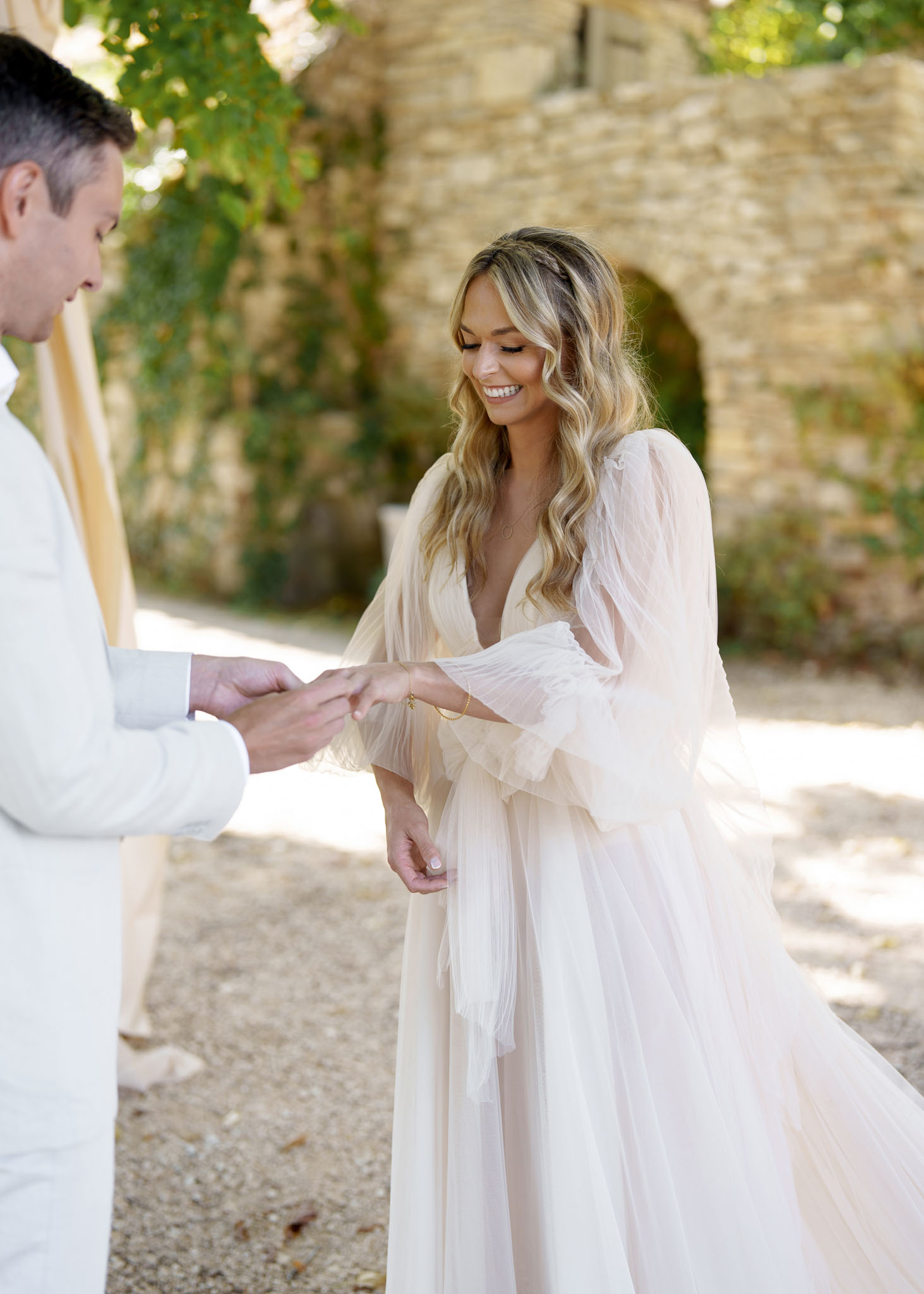 Groom in white suit placing ring on bride in blush tulle gown at outdoor stone courtyard ceremony