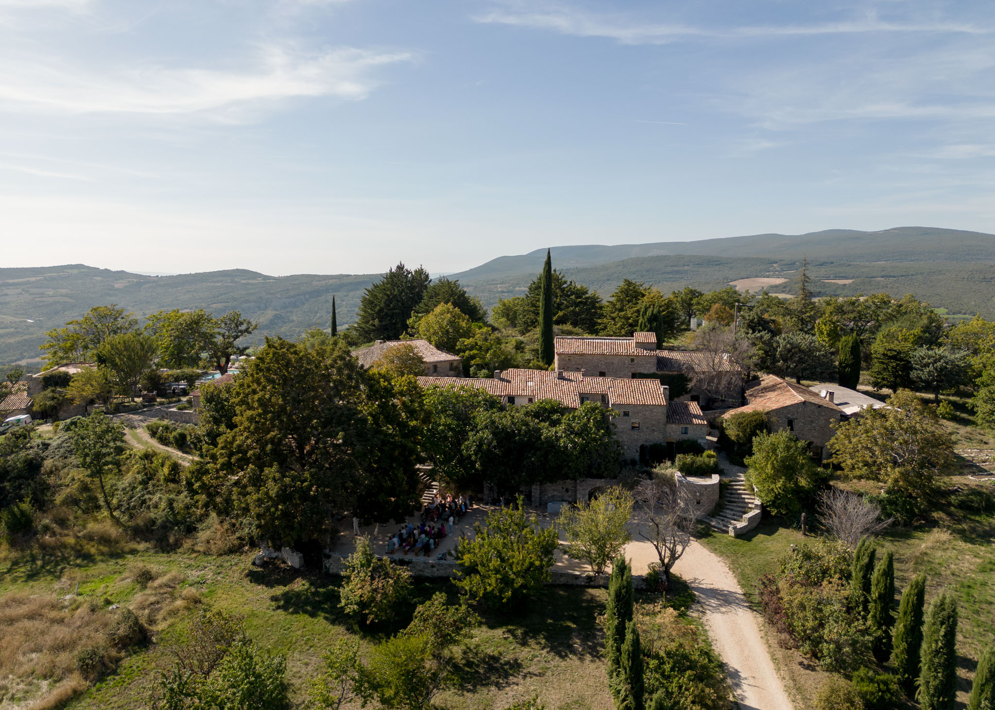 An aerial wide shot of a traditional Provençal stone farmhouse complex with terracotta-tiled roofs, set on an elevated hillside position with a gravel driveway curving through the grounds. A small gathering of approximately 20–30 guests is visible in an outdoor courtyard area at the lower-left section of the property, suggesting an outdoor ceremony or cocktail moment in progress. The estate features multiple stone outbuildings, cypress trees, and stone staircases connecting different levels of the grounds. The surrounding landscape includes rolling hills and distant mountain ridges. Potential venue feature image.
