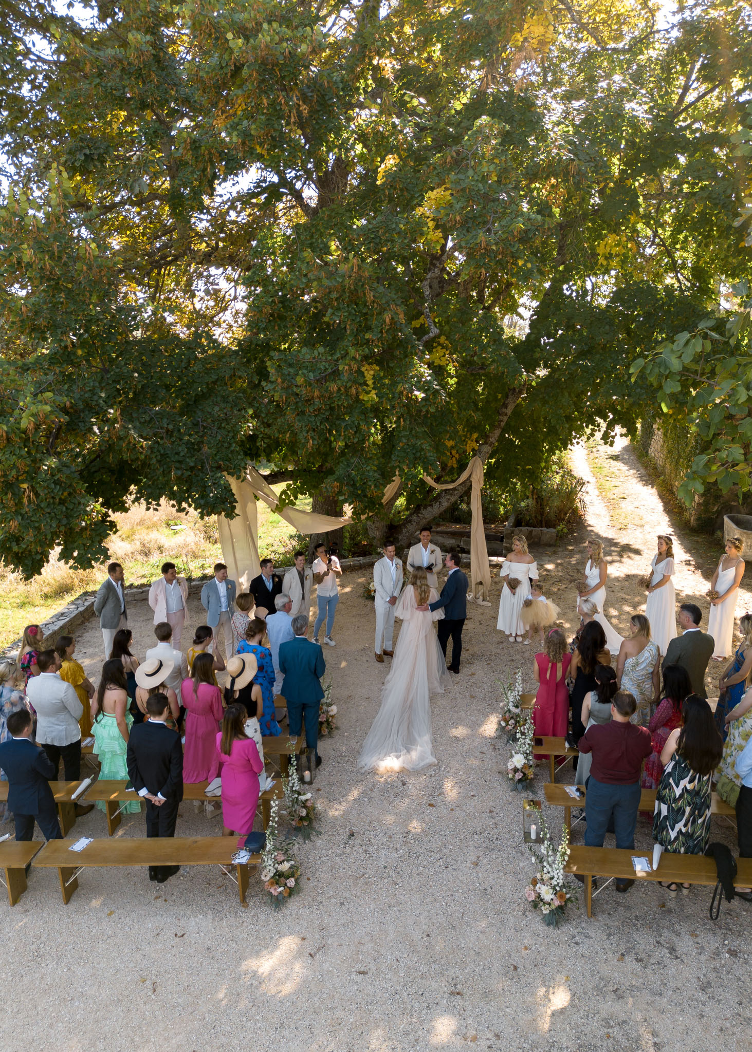 An outdoor wedding ceremony captured from an elevated angle, showing the bride in a blush-pink gown with a long flowing veil walking toward the groom, who is dressed in a dark navy suit, at the altar beneath large mature trees. The ceremony space is set on a gravel surface with wooden benches arranged in two rows on either side of the aisle, decorated with low floral arrangements featuring white delphiniums, blush roses, and greenery at the bench ends. A sandy-toned draped fabric arch frames the altar area, where a groomsman group in light beige and grey suits stands to one side, and bridesmaids in white off-the-shoulder dresses stand to the other. Approximately 50 guests are standing and watching the ceremony, dressed in a mix of colorful outfits including fuchsia, cobalt blue, mustard yellow, and mint green, giving the gathering a relaxed, warm-weather feel. The overall styling is rustic and natural, with a neutral and earthy decor palette accented by the warm afternoon light filtering through the tree canopy.
