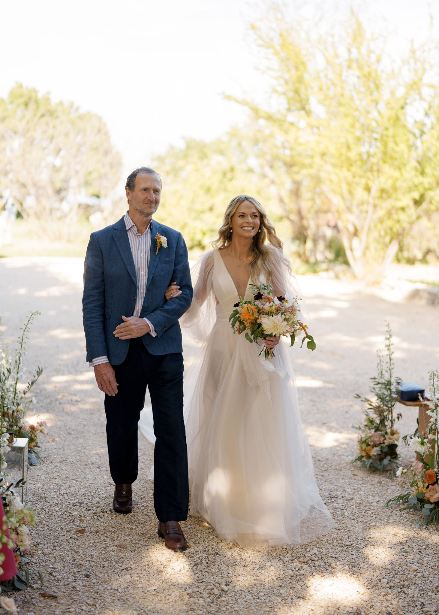 Bride in tulle gown with sheer sleeves walking down a flower-lined gravel aisle with her father in natural daylight