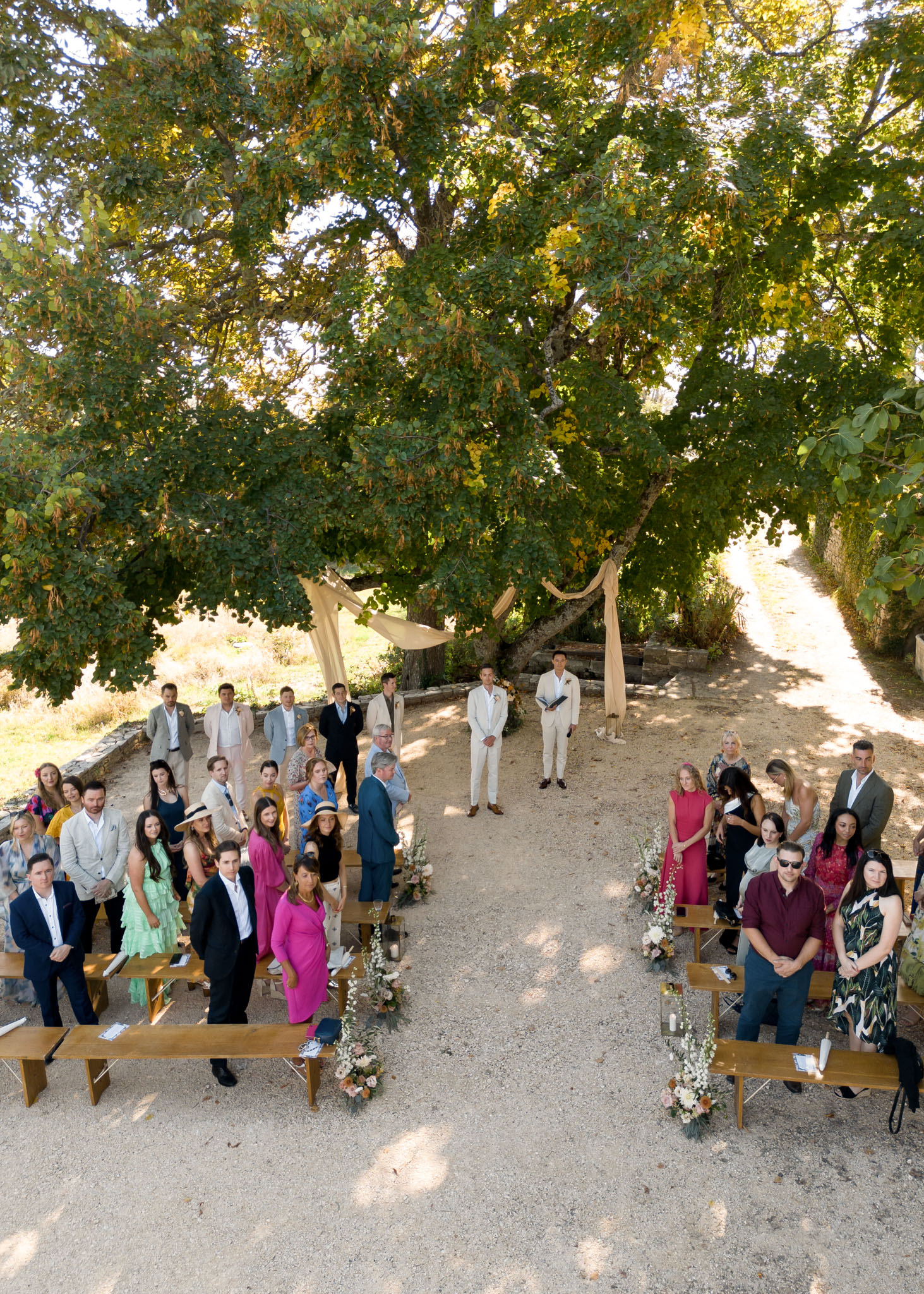 An outdoor wedding ceremony photographed from an elevated angle, showing approximately 40–50 guests seated and standing on wooden benches arranged in two sections with a central gravel aisle. The ceremony is taking place beneath a large mature tree, with the groom in an ivory suit and an officiant in a dark suit standing at the altar area. Two cream or champagne fabric drapes are tied to the tree branches, framing the altar. Floral arrangements in white, peach, and blush tones are placed along the aisle at the base of the benches, with small glass lanterns interspersed. Guests are dressed in a range of colorful outfits including fuchsia pink, mint green, royal blue, and burgundy. The setting is a rustic outdoor space with a gravel ground and low stone border walls visible in the background, suggesting a countryside or domaine venue. The wide overhead shot captures the full ceremony layout and surrounding landscape.
