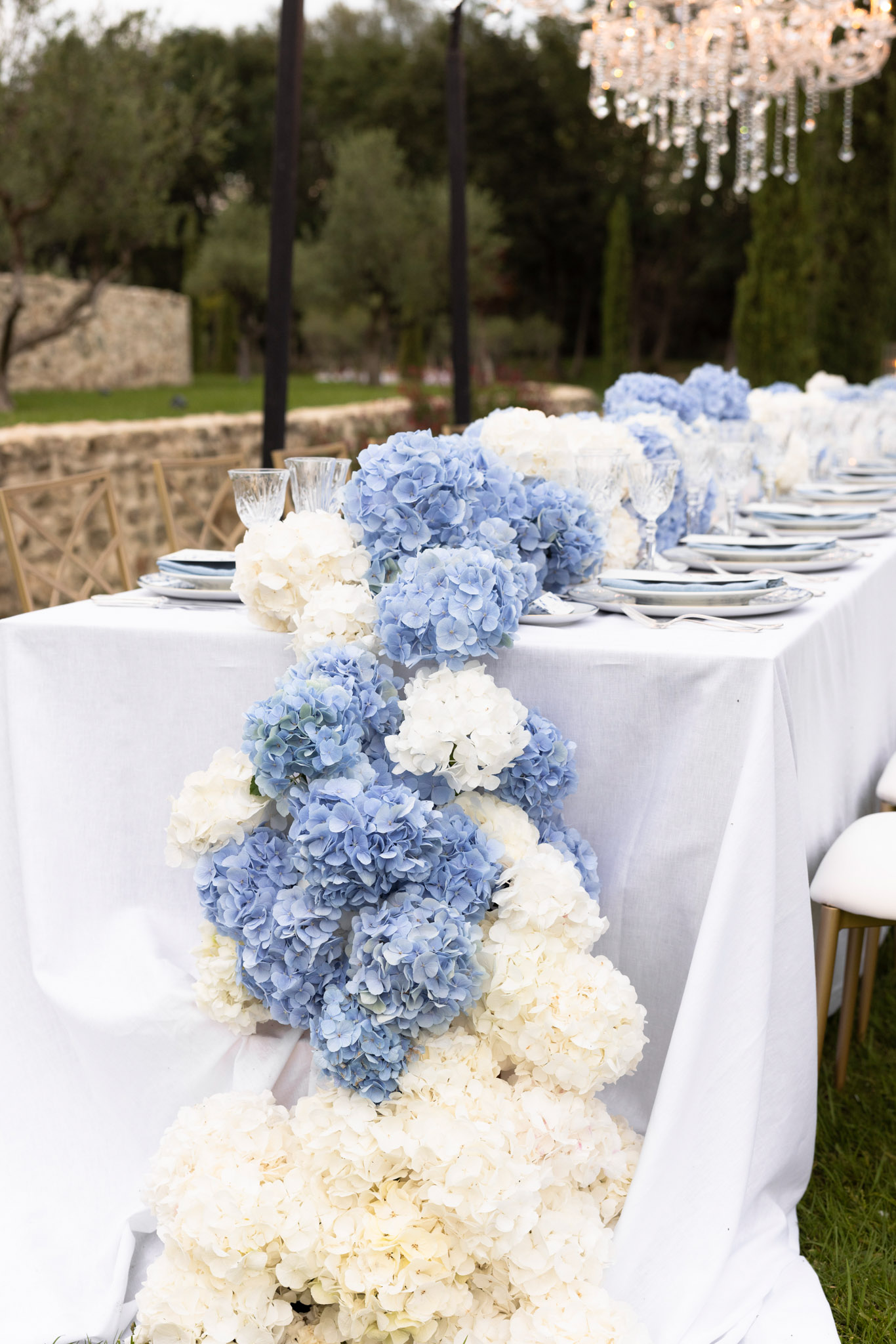 A close-up detail shot of an outdoor wedding reception table set in a garden setting. The long rectangular table is dressed in a white linen tablecloth and features a dense floral runner composed entirely of periwinkle blue and ivory white hydrangeas, cascading down the full length of the table and spilling over the end closest to the camera. Place settings include stacked white and grey-rimmed plates, crystal glassware, and grey linen napkins. Gold chairs are partially visible at the sides. A crystal chandelier hangs in the upper right corner, indicating overhead lighting installation above the outdoor dining space. The overall decor palette is blue, white, and gold, with a classic formal styling approach.