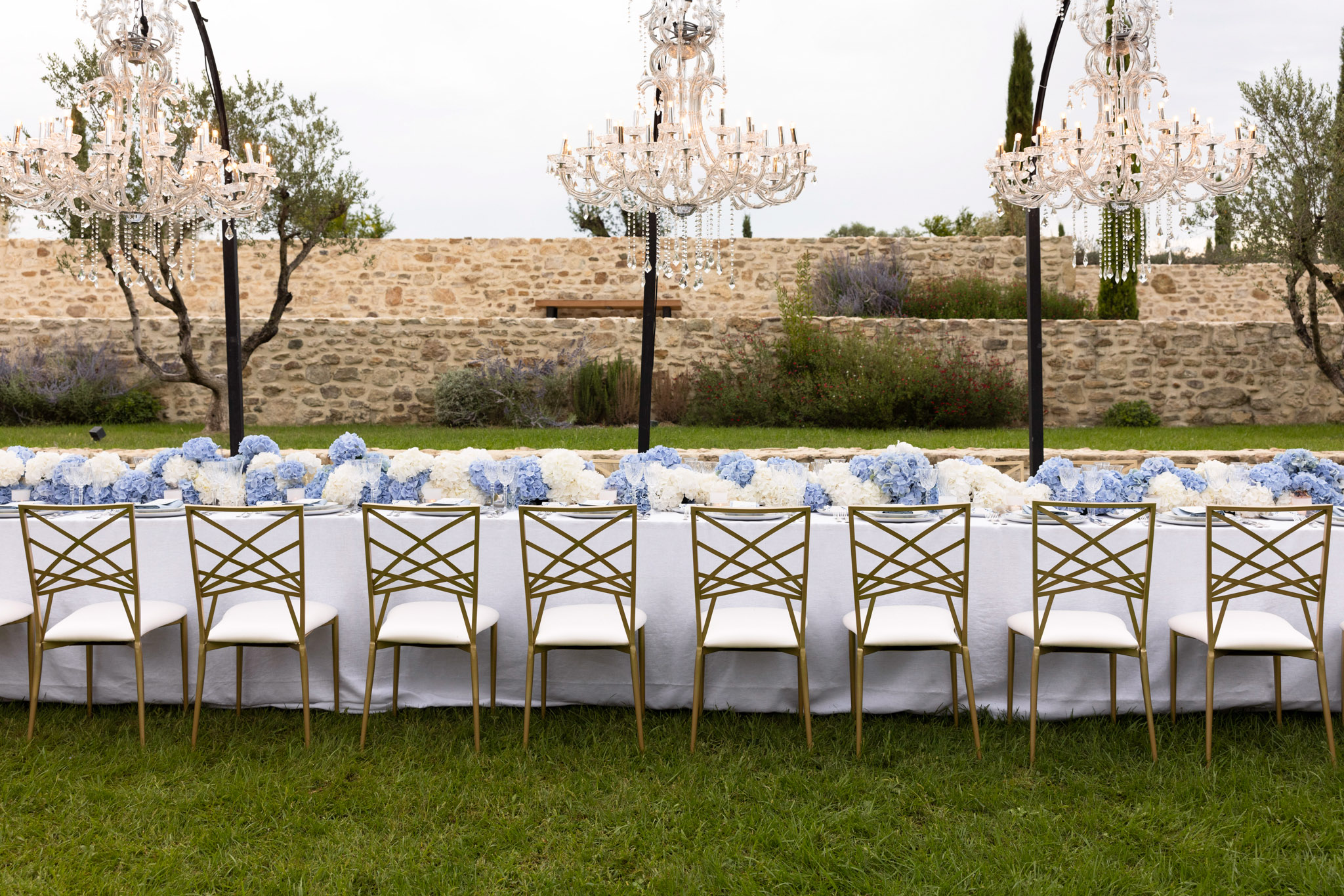 Long banquet table with blue and white hydrangea runner, gold cross-back chairs, and crystal chandeliers in walled garden