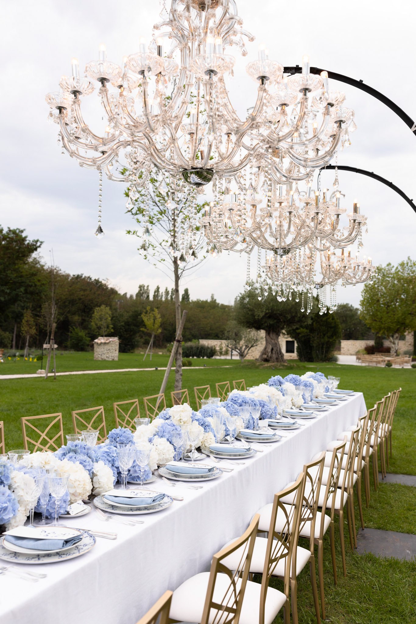Banquet table with blue hydrangea runner gold cross-back chairs and crystal chandeliers on arc frames