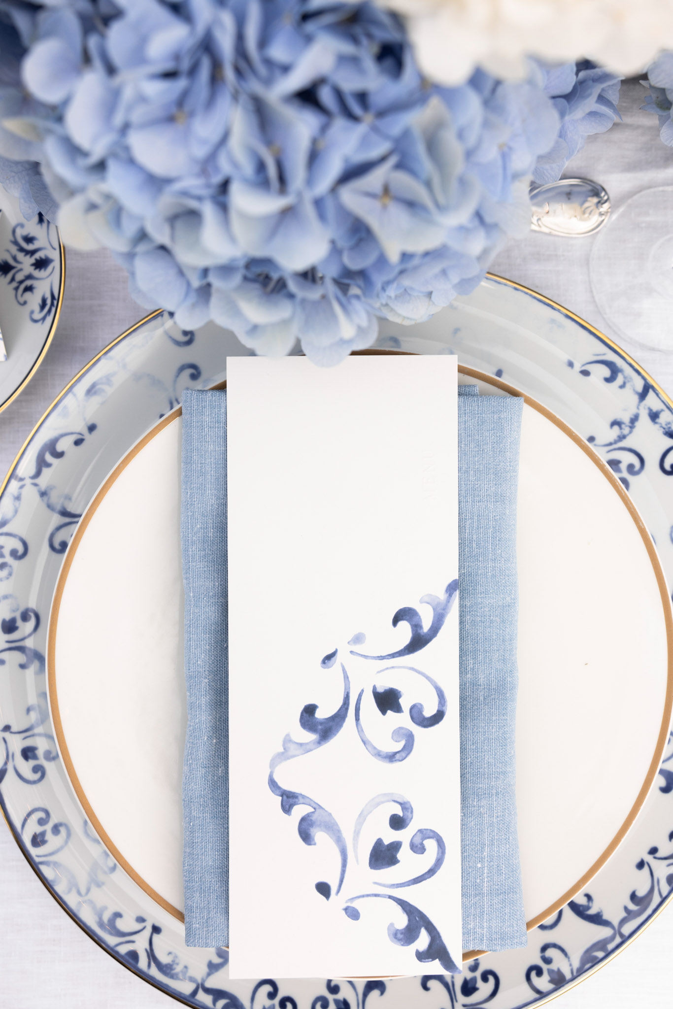 Wedding place setting with navy-bordered charger plate, blue napkin, watercolor menu card, and hydrangea centerpiece