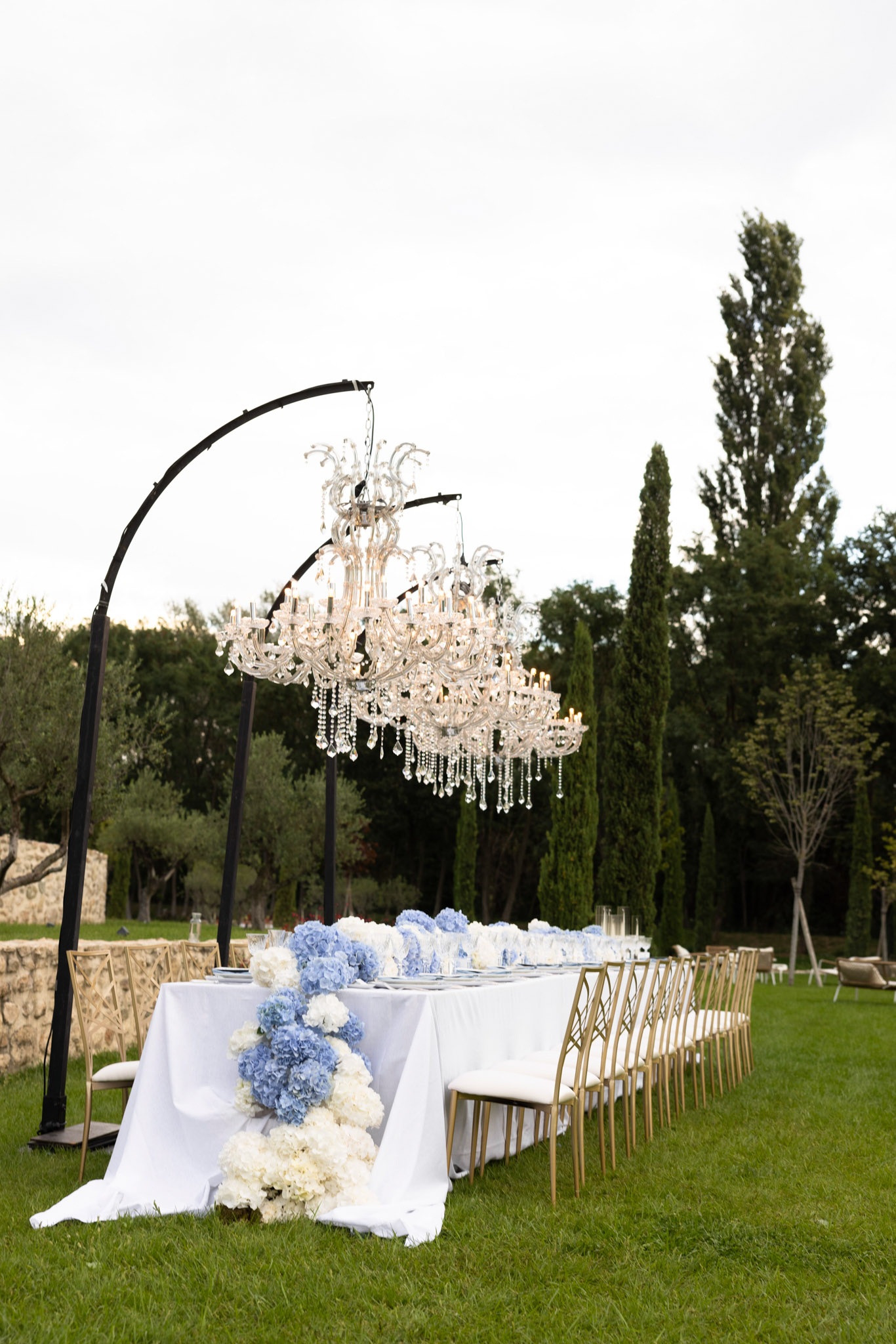 An outdoor wedding reception table setup on a manicured lawn, featuring a long rectangular table dressed in a white linen tablecloth with a floral runner of blue hydrangeas and white hydrangeas cascading along the center. Gold cross-back chairs line both sides of the table, which is set with glassware and place settings in a blue and white palette. A large crystal chandelier with candle-style arms and hanging crystal drops is suspended from a black curved arc stand positioned at the head of the table, serving as a dramatic overhead focal point. The overall styling is classic and formal, with a blue and white color palette, wide shot taken from the end of the table looking down its length.