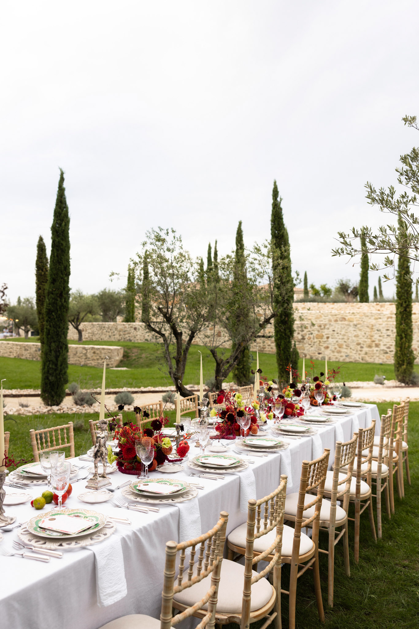 Long table with deep red dahlia and orange centerpieces, botanical china, and cream tapers in garden