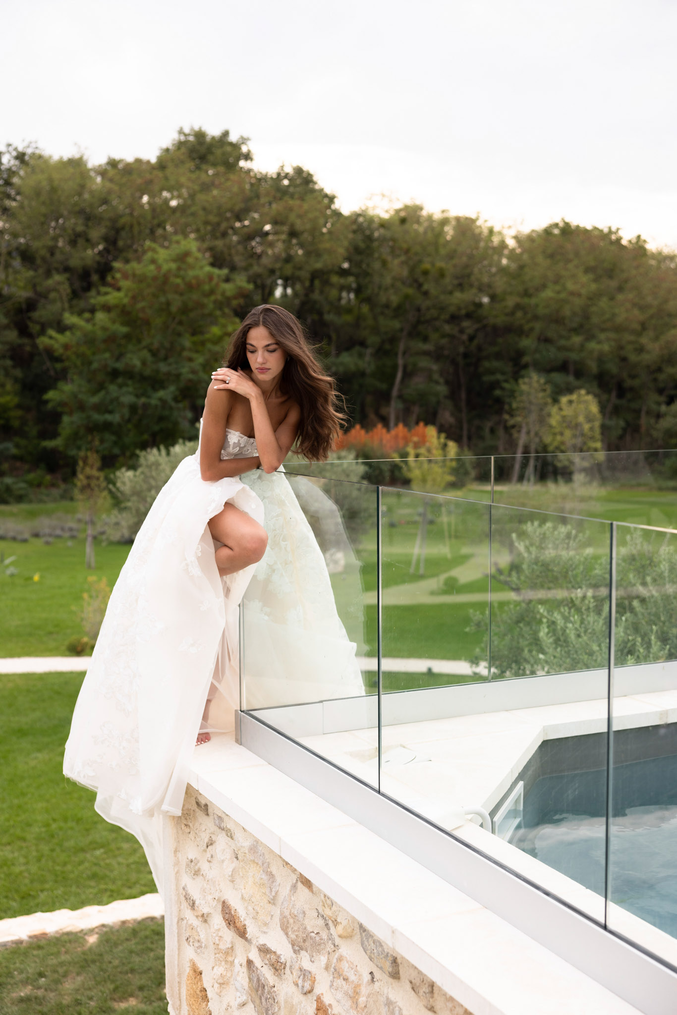 A bridal portrait of a single bride posing on the edge of a contemporary glass-railed terrace or pool deck, leaning against the glass balustrade with her arms folded beneath her chin. She wears a strapless white ballgown with a lace-embellished bodice and a voluminous white overskirt that billows around her, revealing a fitted nude underskirt beneath. Her long dark wavy hair falls loosely over her shoulders. The setting combines rustic stone construction with modern architectural elements — a glass panel railing and a clean white-edged infinity pool or jacuzzi visible below. The surrounding grounds feature a manicured lawn with planted borders, viewed at dusk with soft, fading natural light. The composition is a mid-shot taken from a slightly lower angle, emphasizing the drama of the dress and the modern-meets-stone venue aesthetic. Potential venue feature image.
