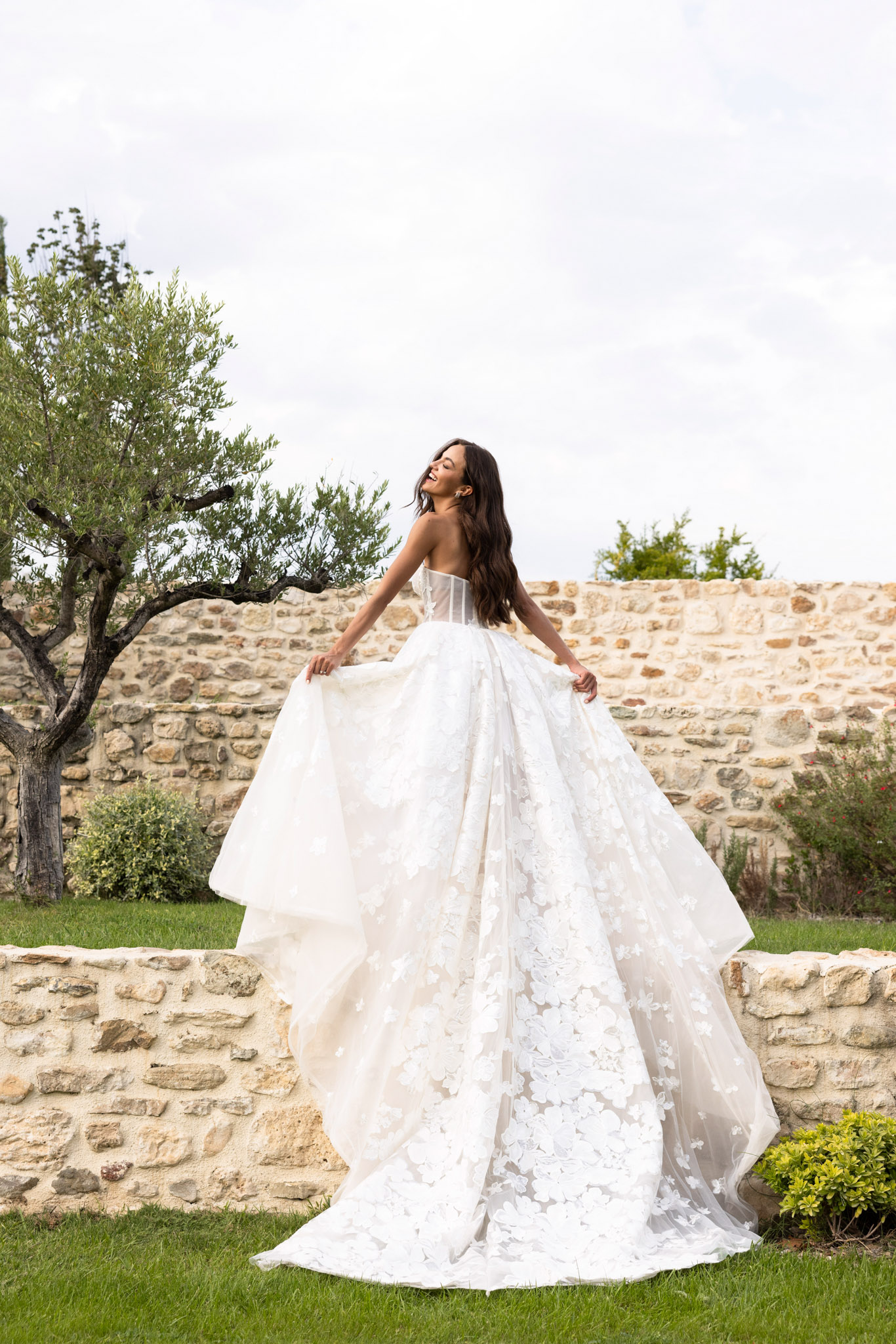 Bride lifting skirt of ivory floral applique ball gown in garden against rustic stone wall