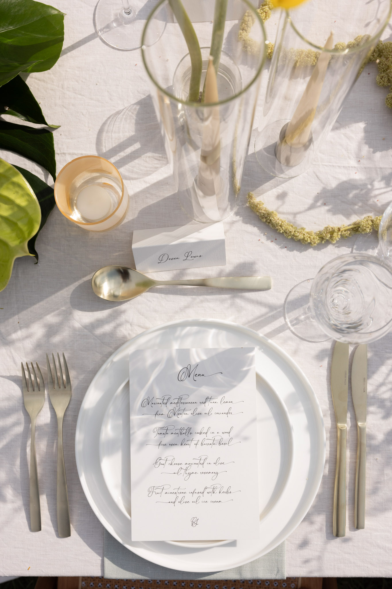 A close-up flat lay detail shot of a wedding reception place setting on a white linen tablecloth, photographed in bright natural sunlight. The place setting features stacked white plates with a calligraphy menu card on top listing four courses in script handwriting, flanked by matte silver cutlery including two forks on the left and a knife on the right, with a matte gold-rimmed votive candle holder and clear glassware including a wine glass and water glass. A handwritten calligraphy place card reading 'Devon Lewis' sits above the plate alongside a matte gold spoon. The centerpiece styling includes tall clear cylindrical vases holding long-stemmed greenery and what appears to be dried yellow flowers, with dried chartreuse floral garland and large tropical green leaves visible at the upper left. The overall palette is white, silver, and gold with natural green accents, reflecting a modern minimalist styling approach.