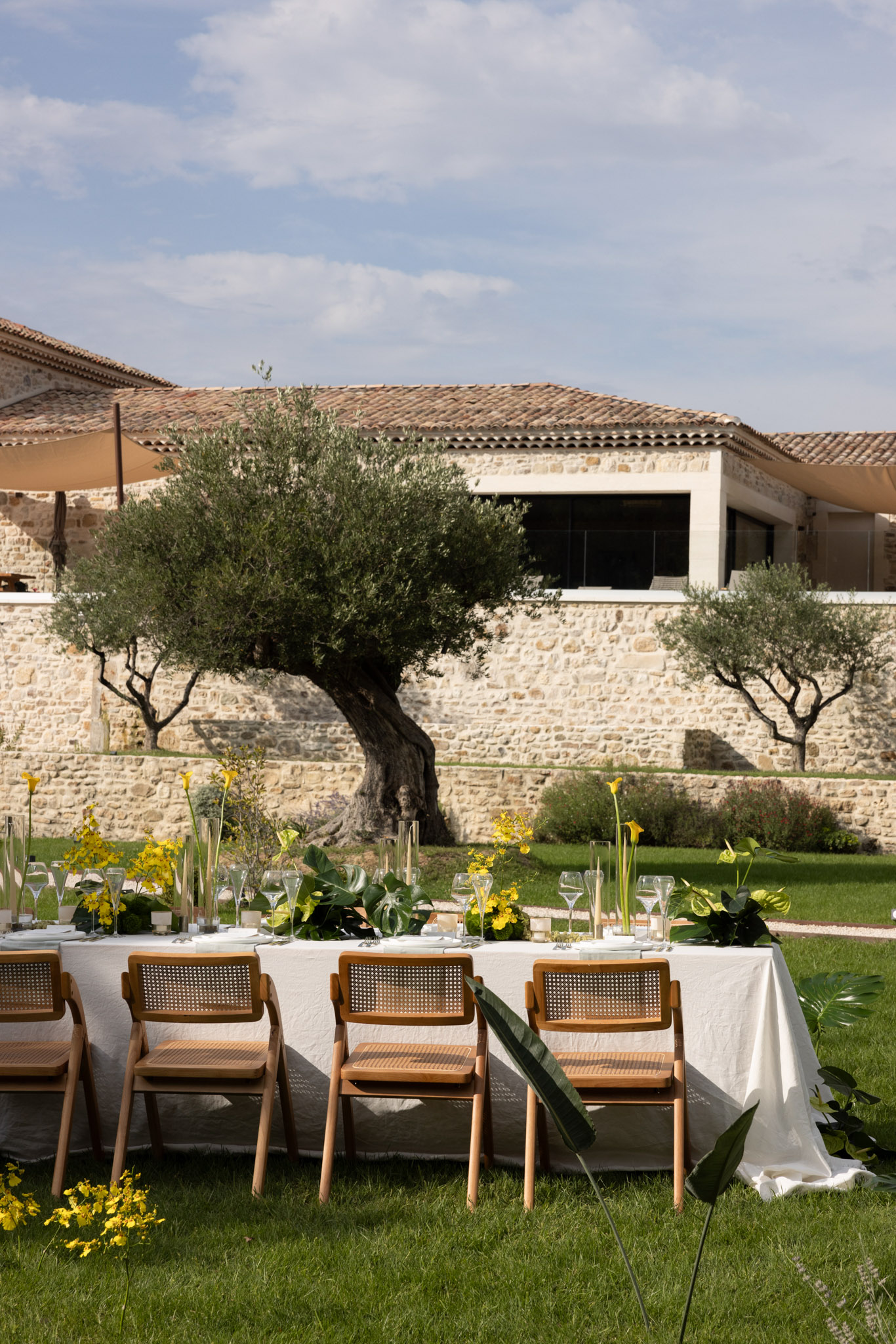 Long table with tropical foliage runner, yellow calla lilies, and rattan chairs before stone property