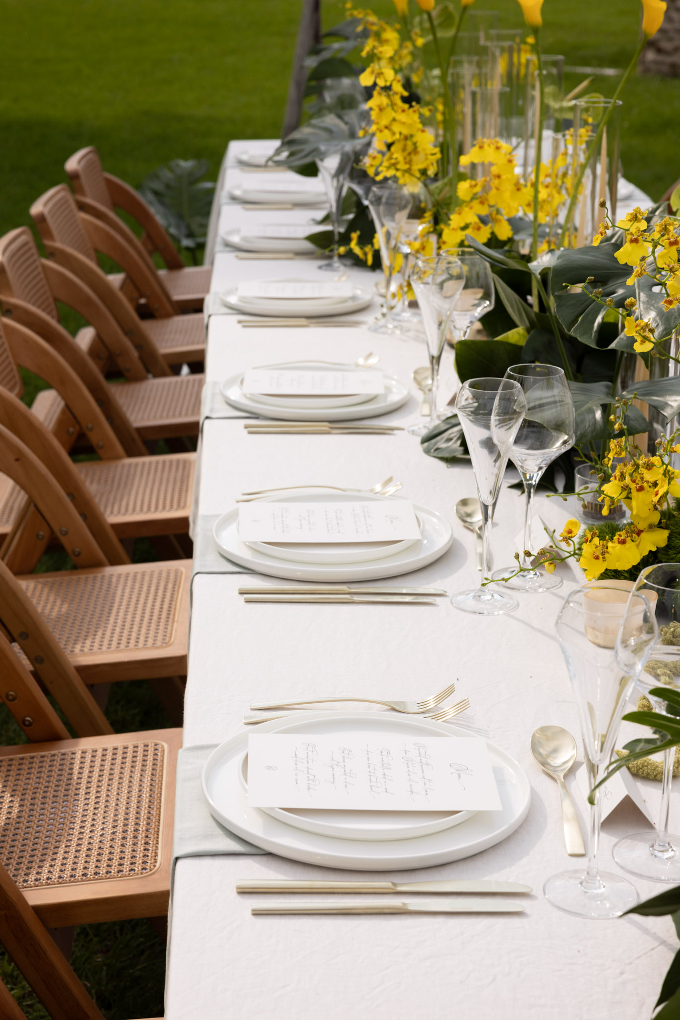 Reception table with yellow oncidium orchids and tropical leaves centerpiece, gold flatware, and cane-back chairs