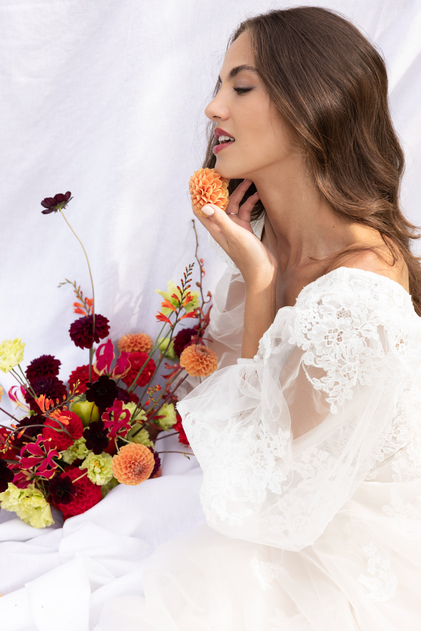 Bride in profile holding orange dahlia beside bouquet of burgundy dahlias and gloriosa lilies