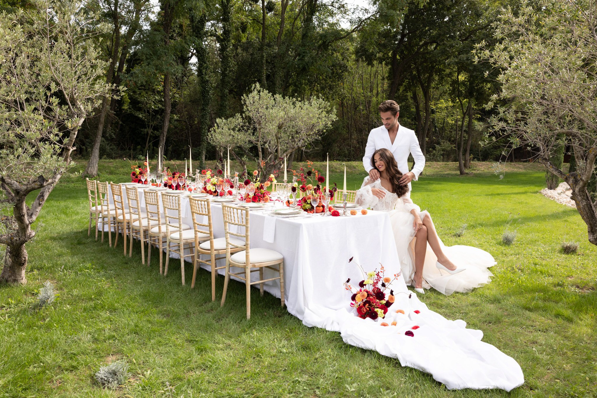 A couple poses outdoors beside a long rectangular reception table set in a garden orchard with olive trees. The bride is seated at the end of the table wearing a long-sleeved lace corset wedding gown with a voluminous train that extends across the lawn, while the groom stands behind her in an all-white suit. The table is dressed in a white linen cloth and lined with gold chiavari chairs with ivory cushions; it is styled with tall ivory taper candles in silver candleholders, gray charger plates, crystal glassware, and bold floral centerpieces in deep red, burgundy, orange, chartreuse, and coral — featuring dahlias and ranunculus. A loose floral cluster of the same red, burgundy, and orange blooms sits on the trailing skirt at the base of the table. Wide-angle portrait shot capturing the full length of the table and surrounding landscape.