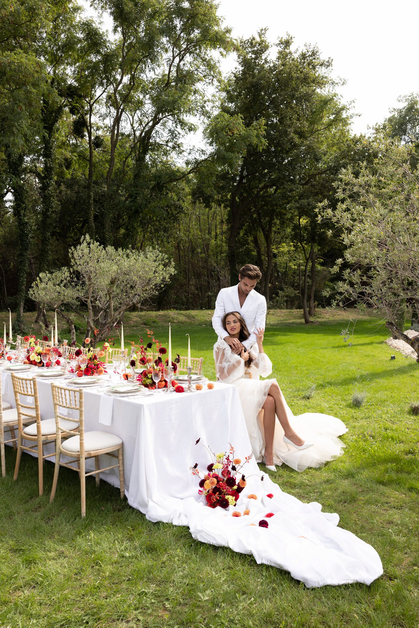 A couple poses beside a long outdoor reception table set in a garden. The bride wears a white gown with sheer bishop sleeves and an extended train that pools across the grass, while the groom wears an all-white suit. The table is dressed with a white linen cloth and styled with gold chiavari chairs, white stacked plates, glassware, tall ivory taper candles, and low floral arrangements featuring deep burgundy and red dahlias, orange and yellow blooms, and trailing red foliage. A loose cluster of burgundy, orange, and yellow dahlias is placed on the bride's train at the base of the table. The color palette for the florals is a rich autumn mix of crimson, burnt orange, mustard, and deep red. The shot is a full-length wide portrait taken in natural daylight, with the couple centered against a backdrop of mature trees and a well-maintained lawn.