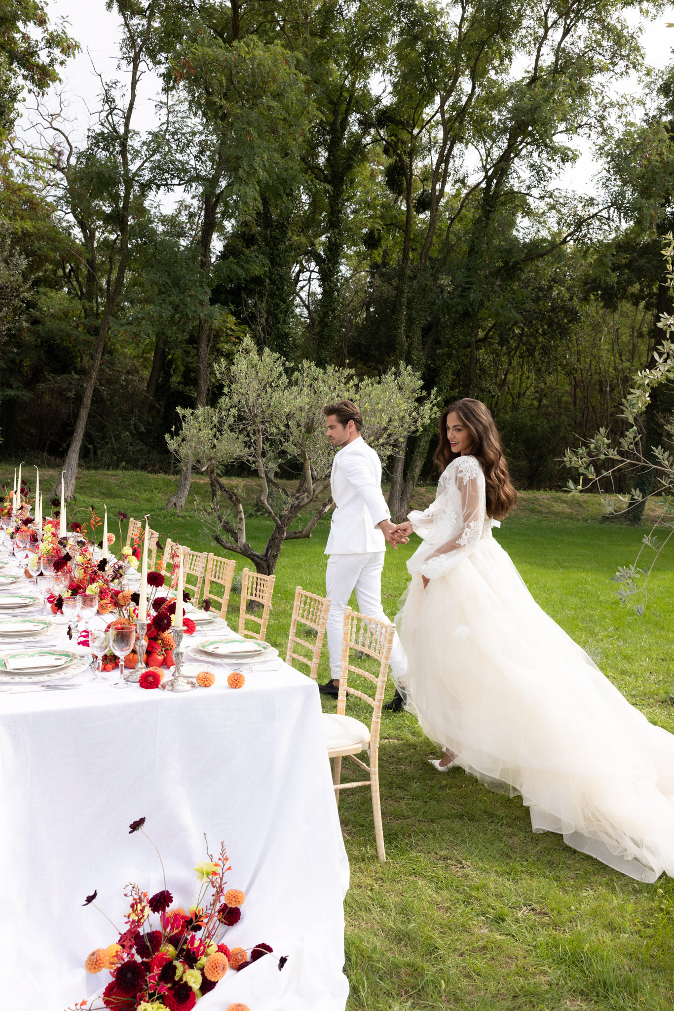 Couple in white beside long reception table with burgundy and orange dahlia runner and taper candles among olive trees