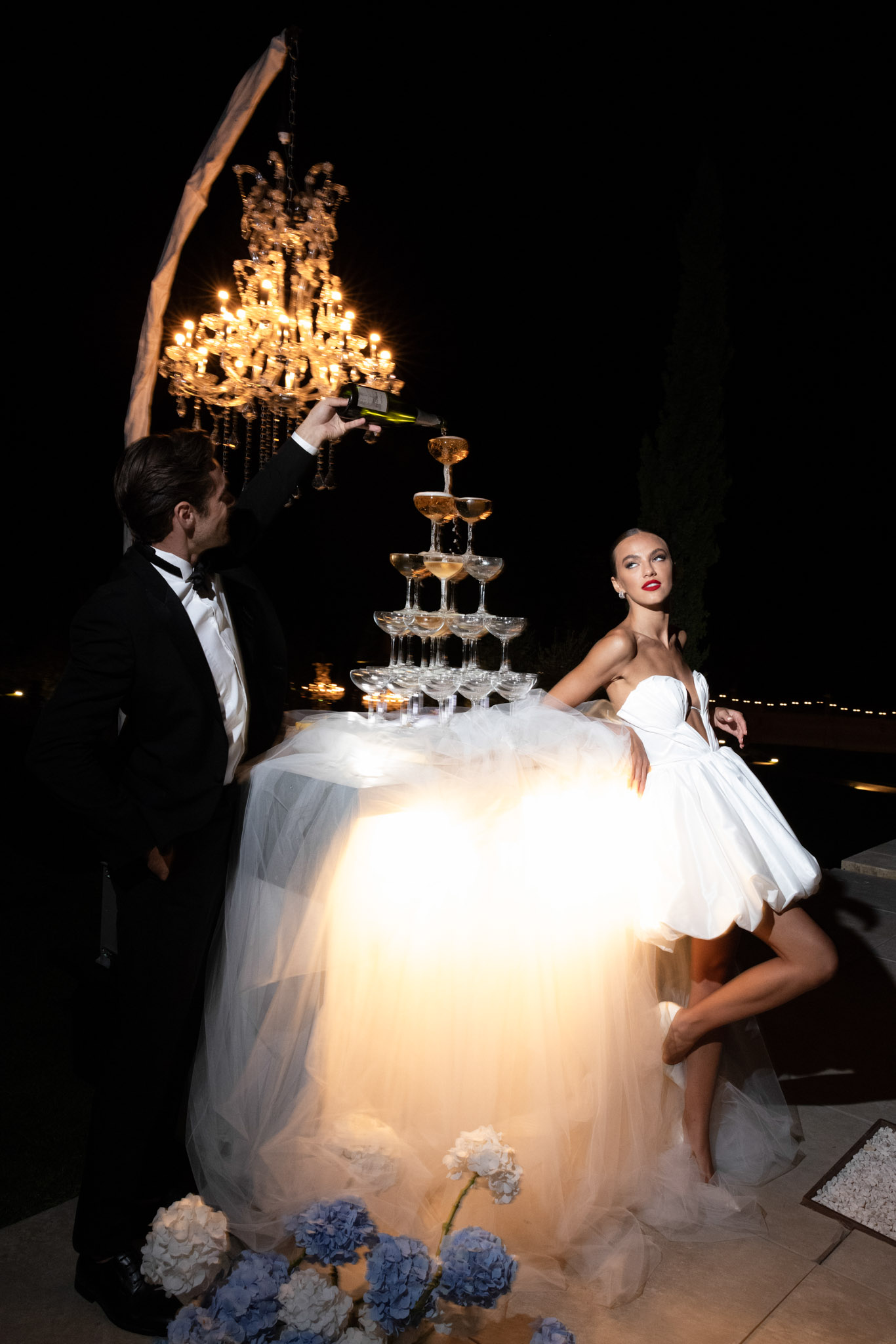 Groom pouring champagne into coupe glass tower while bride in short white tulle gown watches at nighttime reception