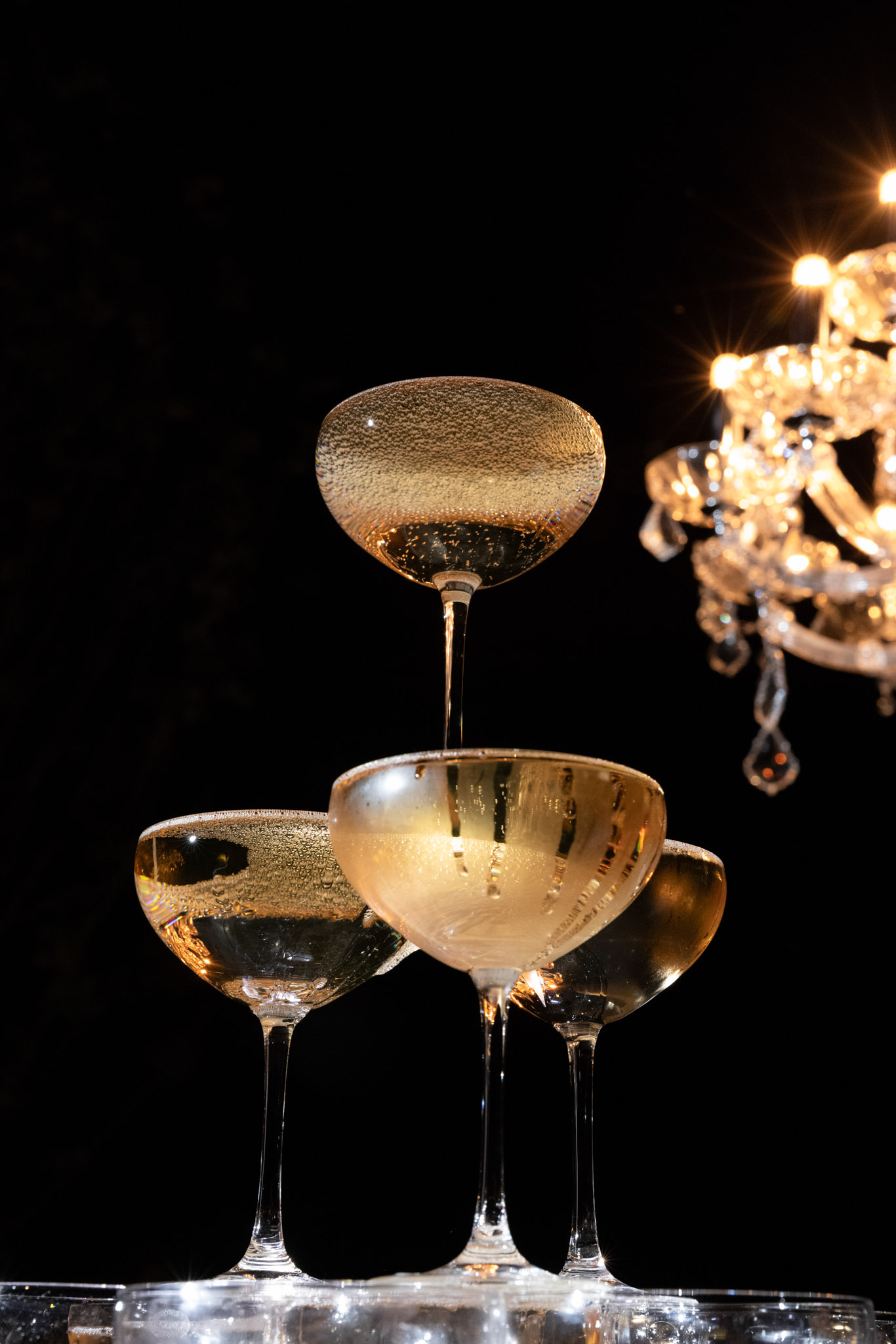 Champagne cascades from top coupe glass down three-tier tower with crystal chandelier glow in dark backdrop