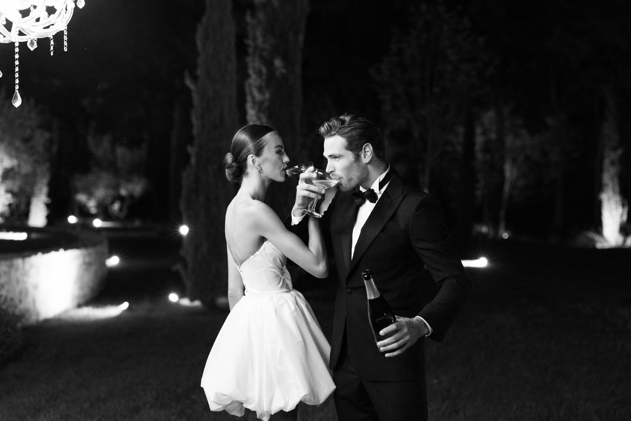 This black-and-white portrait captures a bride and groom sharing a champagne coupe in a crossed-arms toast during an outdoor evening reception. The bride wears a short strapless ballgown-style dress with a voluminous skirt and a low open back, her hair pulled into a sleek low bun; the groom is dressed in a classic black tuxedo with a bow tie and holds a champagne bottle in his free hand. The setting is an outdoor garden or grounds at night, with low pathway lighting and uplighting visible in the background, along with a crystal chandelier partially visible in the upper left corner. The image is shot in high-contrast black and white with deep shadows and bright highlights, giving it a crisp, formal aesthetic; it is a mid-length portrait composition of just the two.