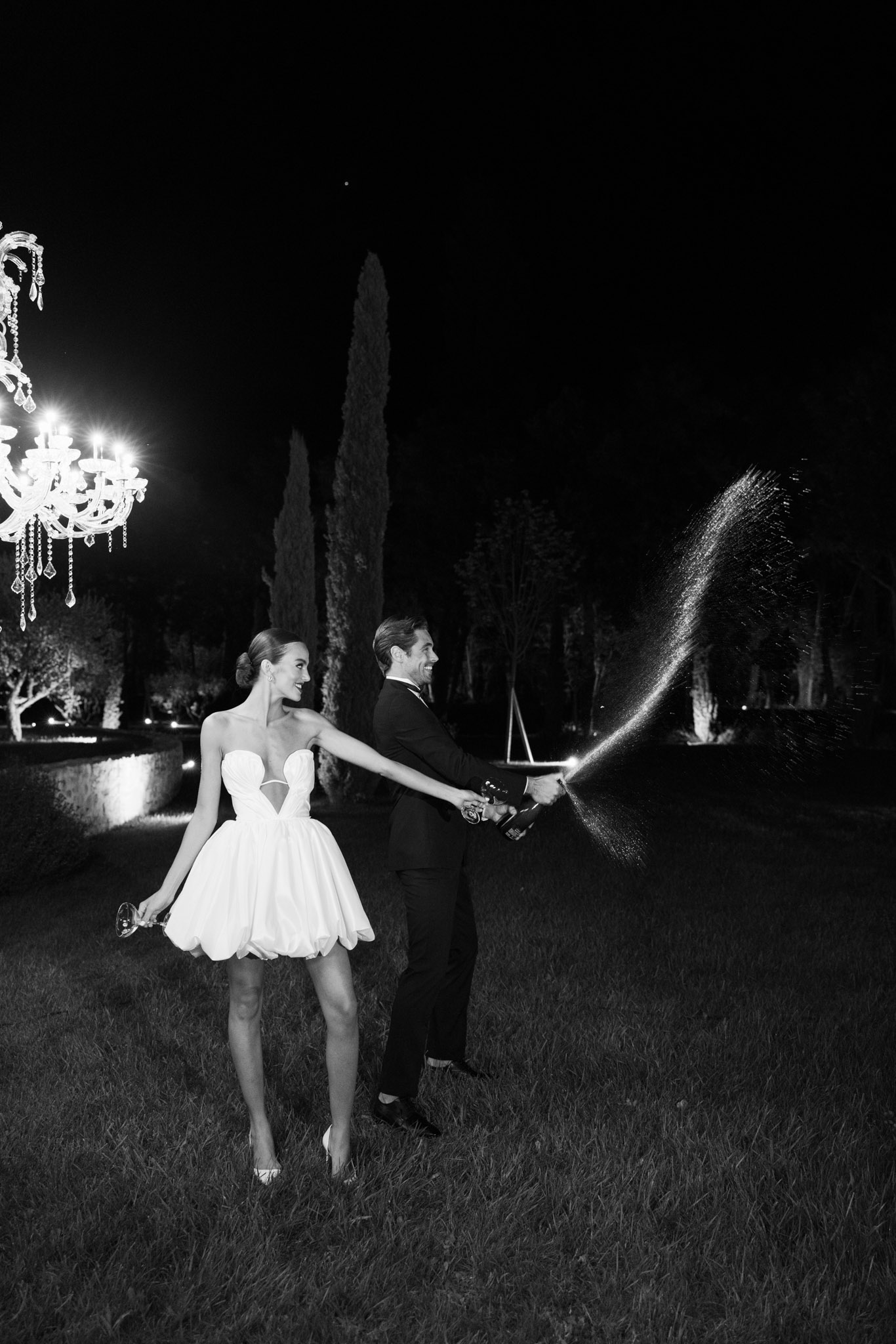 Black and white couple spraying champagne at night with crystal chandelier and cypress uplighting